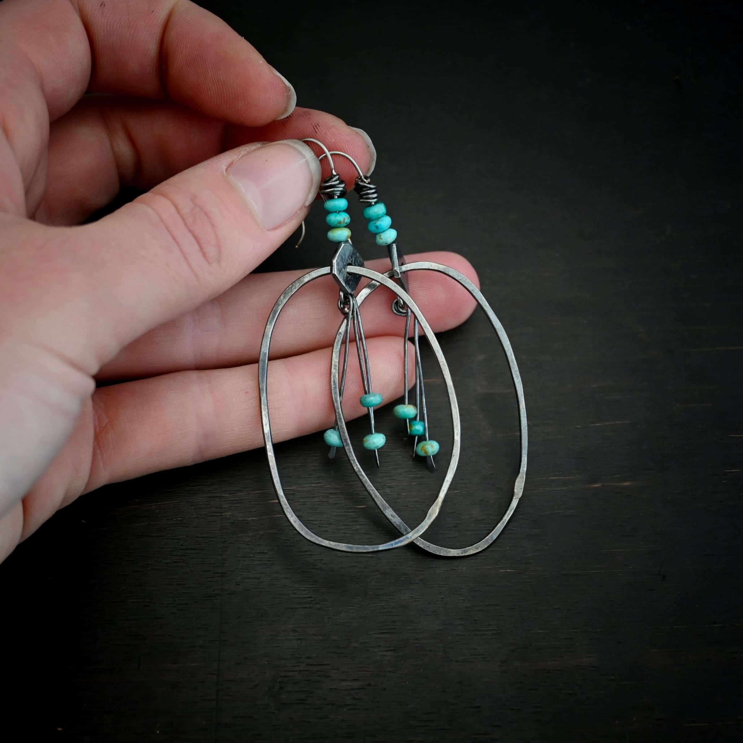 Photo of the "Montana Sky" Earrings. Made out of sterling silver and turquoise. Earrings have french hook ear wires with silver wrapped around the top of them. There are three turquoise beads attached to the top of the ear wires as well. After the turquoise, there is a square, thin piece of silver set sideways with a hole in the center. Going through the hole is a large oval hoop that is hammered flat, but smooth. Hanging from the bottom of the square off of a soldered jump ring, in the center of the oval are three small, thin sticks of silver with loops on the top and hammered paddles on the bottom. Hanging just above the paddles on each stick is a small turquoise bead. Earrings are held in the fingertips of a hand.