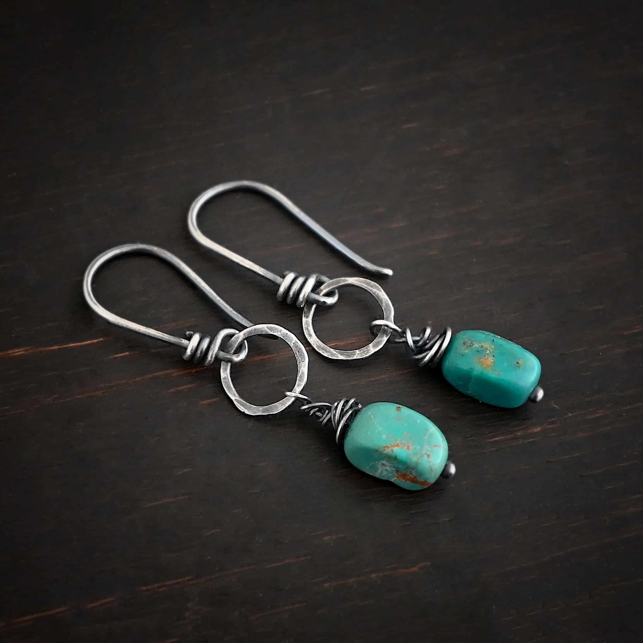 Photo of the "Country Sky" Earrings. Made out of sterling silver and turquoise. Earrings have french hook ear wires with loops on the bottom, and sterling silver wrapped around the bottom before the loop. The loop is attached to a medium sized hammered and oxidized silver circle. Hanging from the bottom of the circle is a silver stick with a loop on the top, silver wrapped around the stick, and a paddle bottom. In between the wrapped silver and the paddle is an oval shaped turquoise. Earrings are lying against a dark background.