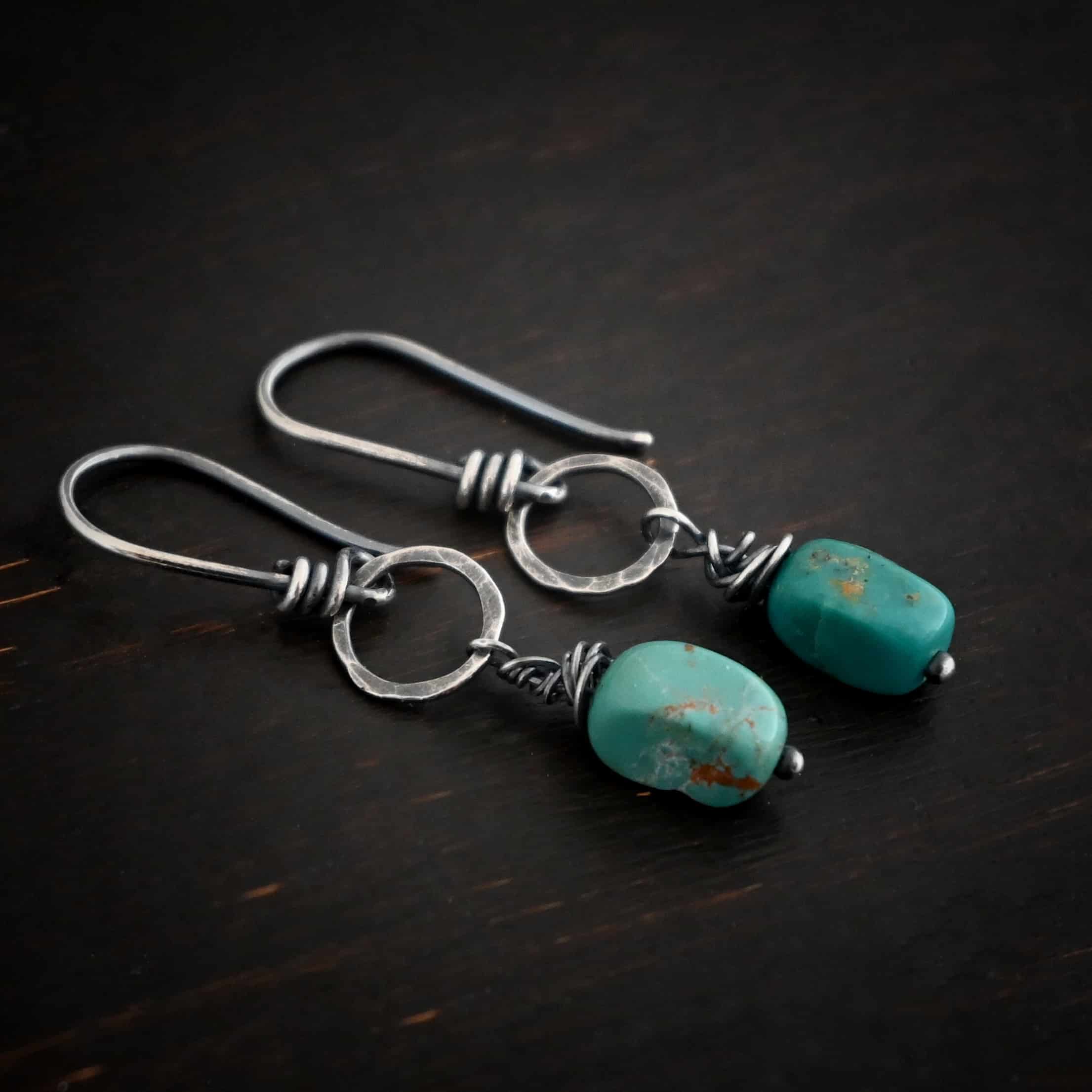 Photo of the "Country Sky" Earrings. Made out of sterling silver and turquoise. Earrings have french hook ear wires with loops on the bottom, and sterling silver wrapped around the bottom before the loop. The loop is attached to a medium sized hammered and oxidized silver circle. Hanging from the bottom of the circle is a silver stick with a loop on the top, silver wrapped around the stick, and a paddle bottom. In between the wrapped silver and the paddle is an oval shaped turquoise. Earrings are lying against a dark background.