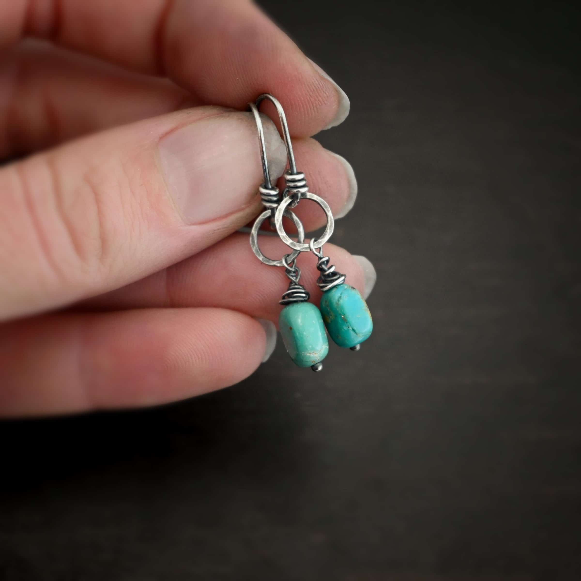 Photo of the "Country Sky" Earrings. Made out of sterling silver and turquoise. Earrings have french hook ear wires with loops on the bottom, and sterling silver wrapped around the bottom before the loop. The loop is attached to a medium sized hammered and oxidized silver circle. Hanging from the bottom of the circle is a silver stick with a loop on the top, silver wrapped around the stick, and a paddle bottom. In between the wrapped silver and the paddle is an oval shaped turquoise. Earrings are held in the fingertips of a hand.