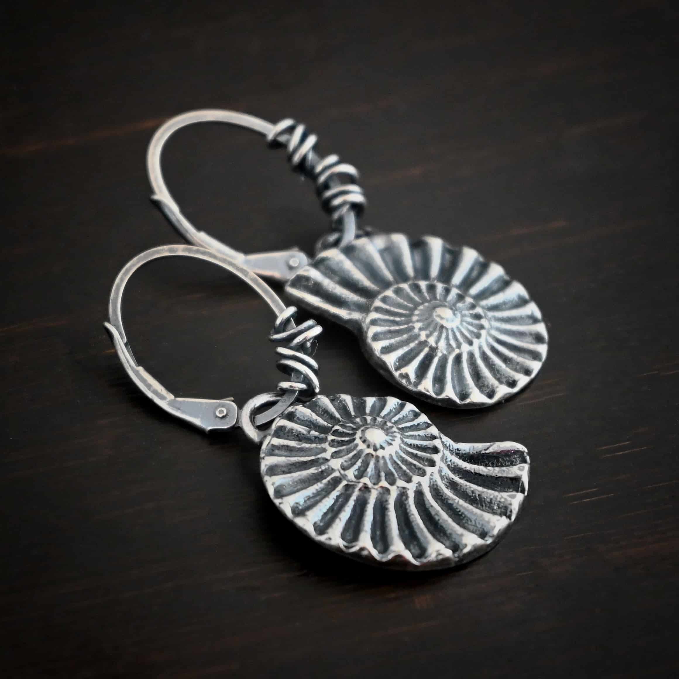 Photo of the "Under The Sea" Earrings. Made out of sterling silver and oxidized. Earrings have leverback ear wires with oxidized silver wrapped around the center part of the ear wires. Attached to the bottom of the ear wires is a loop that is soldered to the top of a casted sterling silver sea shell. The lines of the sea shell are colored in darker to emphasize them. Earrings are lying against a dark background.