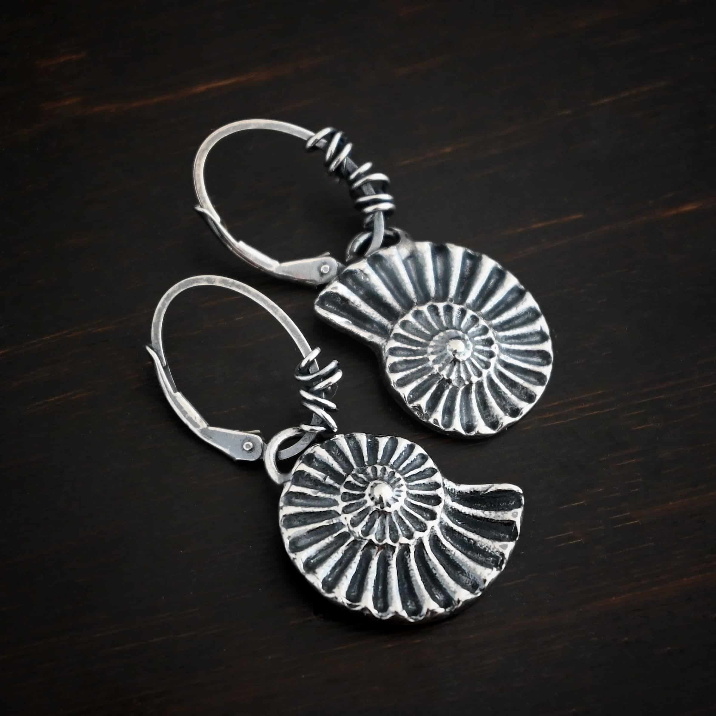 Photo of the "Under The Sea" Earrings. Made out of sterling silver and oxidized. Earrings have leverback ear wires with oxidized silver wrapped around the center part of the ear wires. Attached to the bottom of the ear wires is a loop that is soldered to the top of a casted sterling silver sea shell. The lines of the sea shell are colored in darker to emphasize them. Earrings are lying against a dark background.