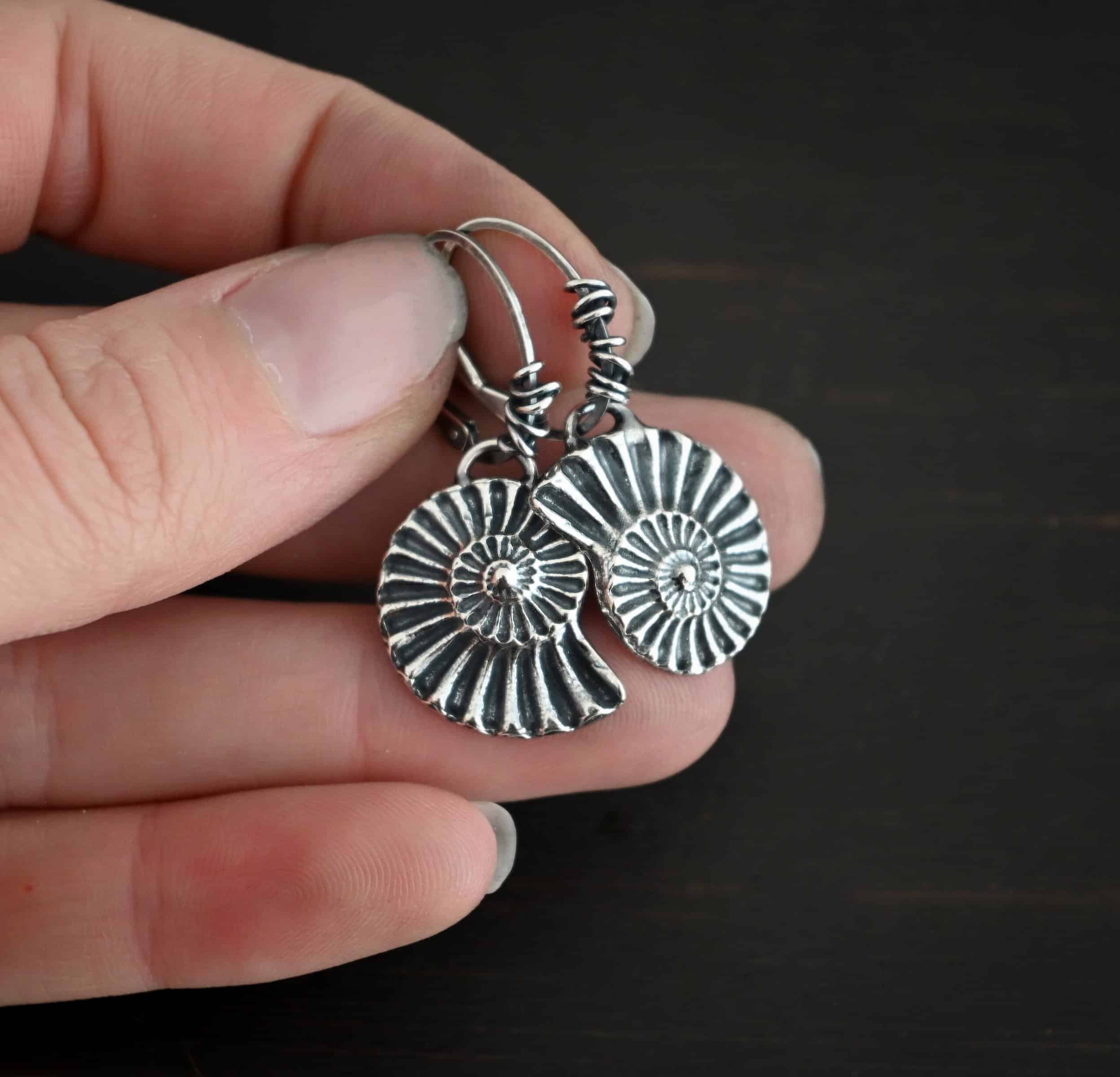 Photo of the "Under The Sea" Earrings. Made out of sterling silver and oxidized. Earrings have leverback ear wires with oxidized silver wrapped around the center part of the ear wires. Attached to the bottom of the ear wires is a loop that is soldered to the top of a casted sterling silver sea shell. The lines of the sea shell are colored in darker to emphasize them. Earrings are held in the fingertips of a hand.