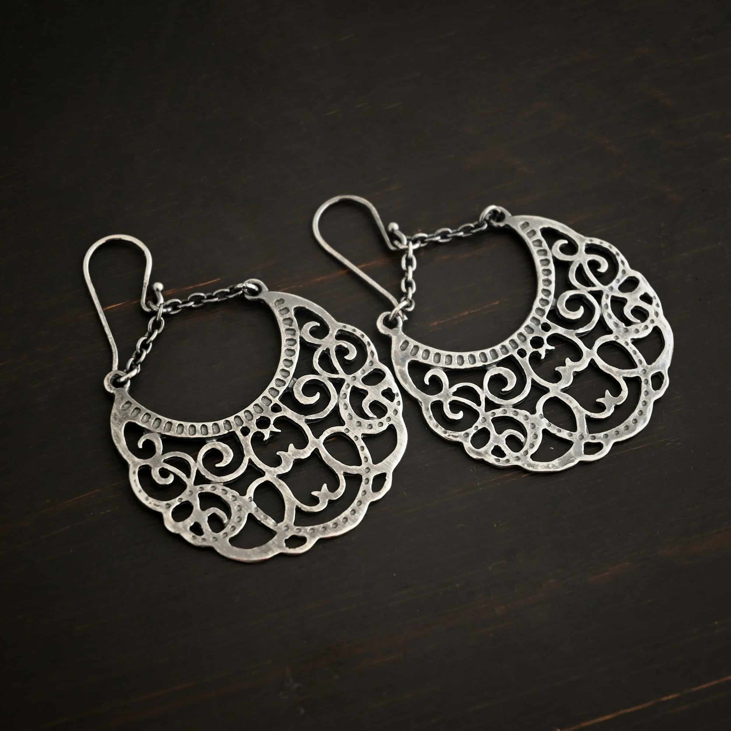 Photo of the "Gypsy Road" Earrings. Made out of sterling silver. Earrings have french hook ear wires with a jump ring attached to them. Attached to the jump ring are two very small strands of sterling silver that go opposite ways to create a long triangle shape. Attached on each side of the strands is a large crescent moon shaped pendant. The insides of the pendant are carved intricately with swirls. The inside curve of the moon has small hammered dots going around it. Earrings are lying against a dark background.