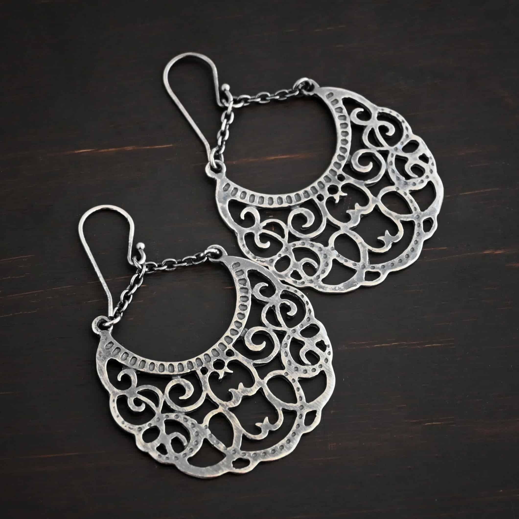 Photo of the "Gypsy Road" Earrings. Made out of sterling silver. Earrings have french hook ear wires with a jump ring attached to them. Attached to the jump ring are two very small strands of sterling silver that go opposite ways to create a long triangle shape. Attached on each side of the strands is a large crescent moon shaped pendant. The insides of the pendant are carved intricately with swirls. The inside curve of the moon has small hammered dots going around it. Earrings are lying against a dark background.