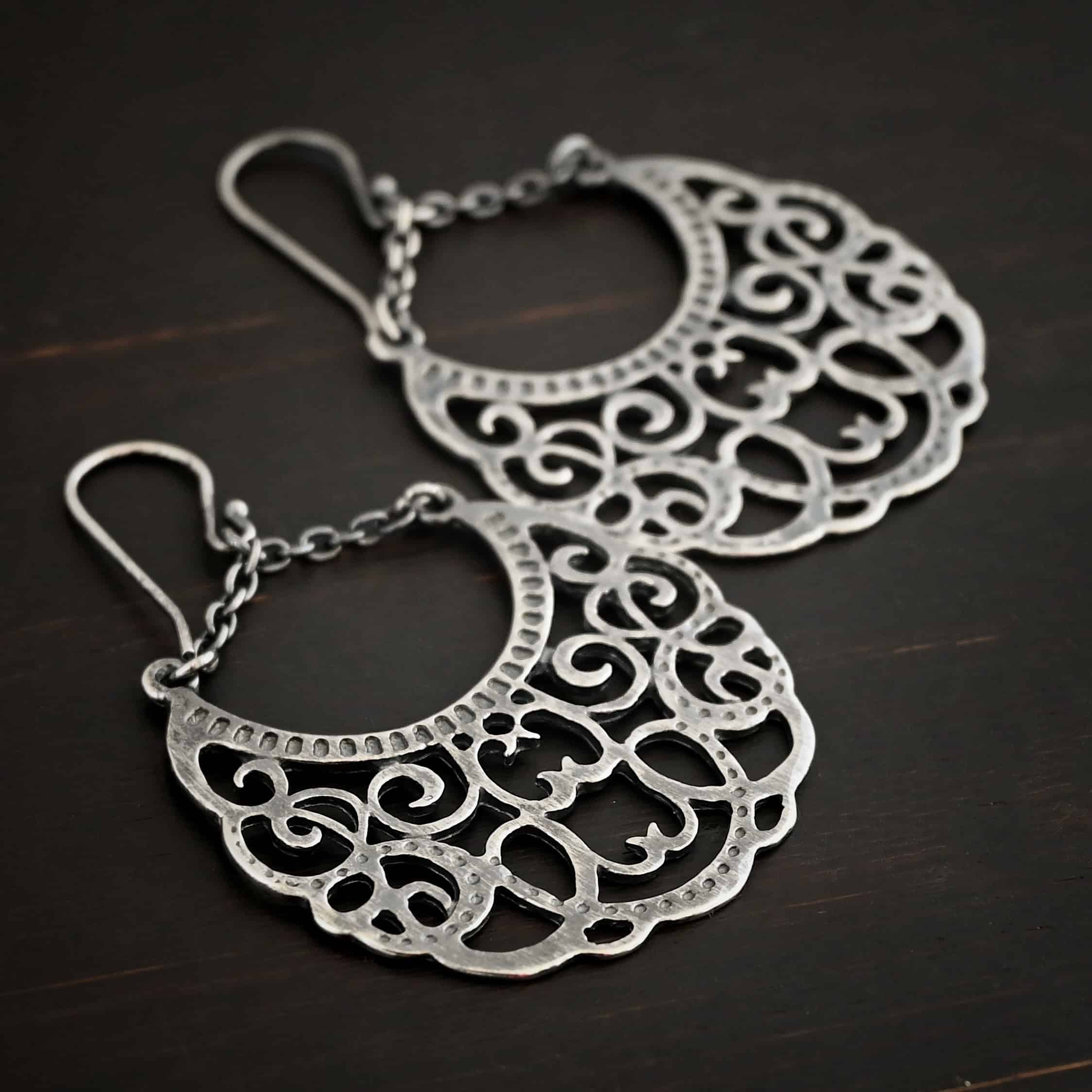Photo of the "Gypsy Road" Earrings. Made out of sterling silver. Earrings have french hook ear wires with a jump ring attached to them. Attached to the jump ring are two very small strands of sterling silver that go opposite ways to create a long triangle shape. Attached on each side of the strands is a large crescent moon shaped pendant. The insides of the pendant are carved intricately with swirls. The inside curve of the moon has small hammered dots going around it. Earrings are lying against a dark background.