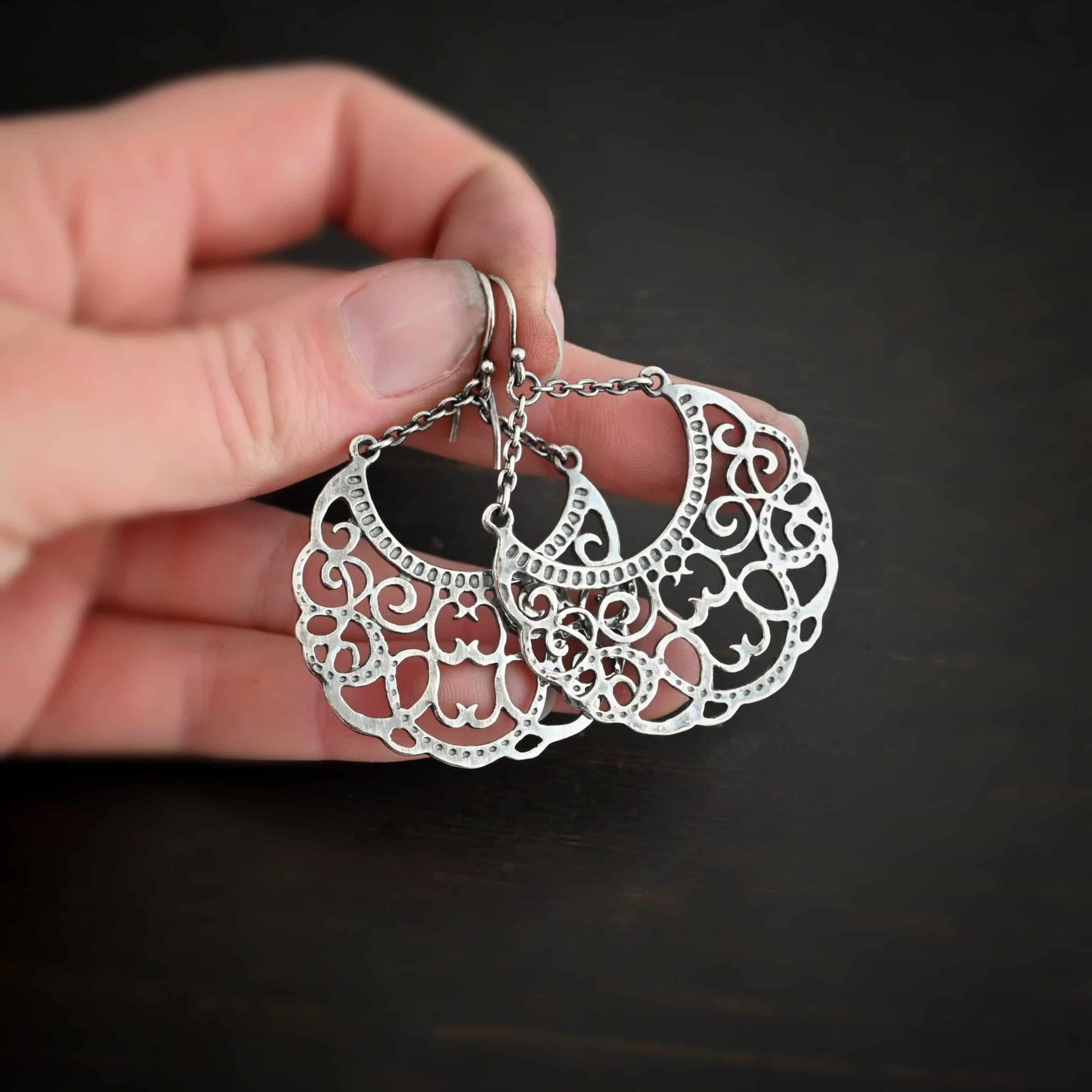 Photo of the "Gypsy Road" Earrings. Made out of sterling silver. Earrings have french hook ear wires with a jump ring attached to them. Attached to the jump ring are two very small strands of sterling silver that go opposite ways to create a long triangle shape. Attached on each side of the strands is a large crescent moon shaped pendant. The insides of the pendant are carved intricately with swirls. The inside curve of the moon has small hammered dots going around it. Earrings are held in the fingertips of a hand.