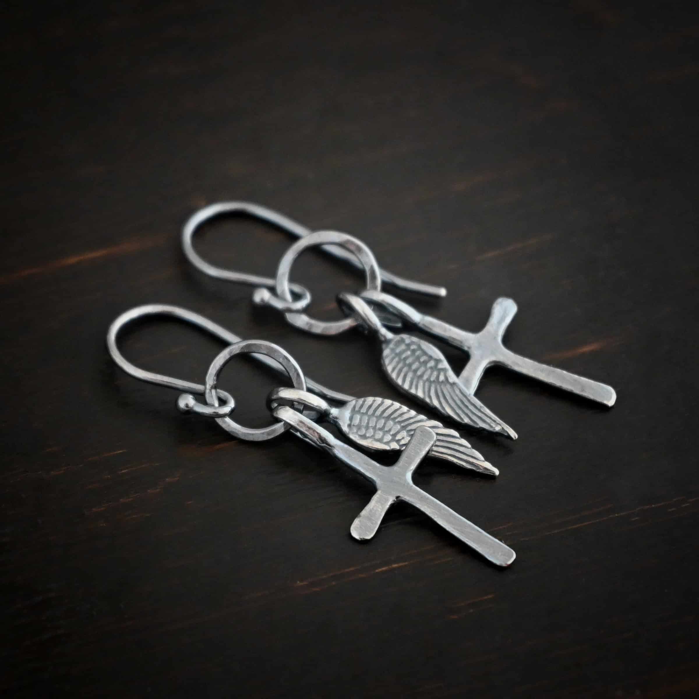 Strength Earrings Photo of the "Strength" Earrings. Made out of oxidized sterling silver. There is a small sized cross that is flat and smooth. It has a loop soldered to the top of it. There is a small sized angel wing pendant next to it, also with a soldered loop on the top. Both of them hang down from a hammered and oxidized circle that hangs on a french hook ear wire. Earrings are lying against a dark background.