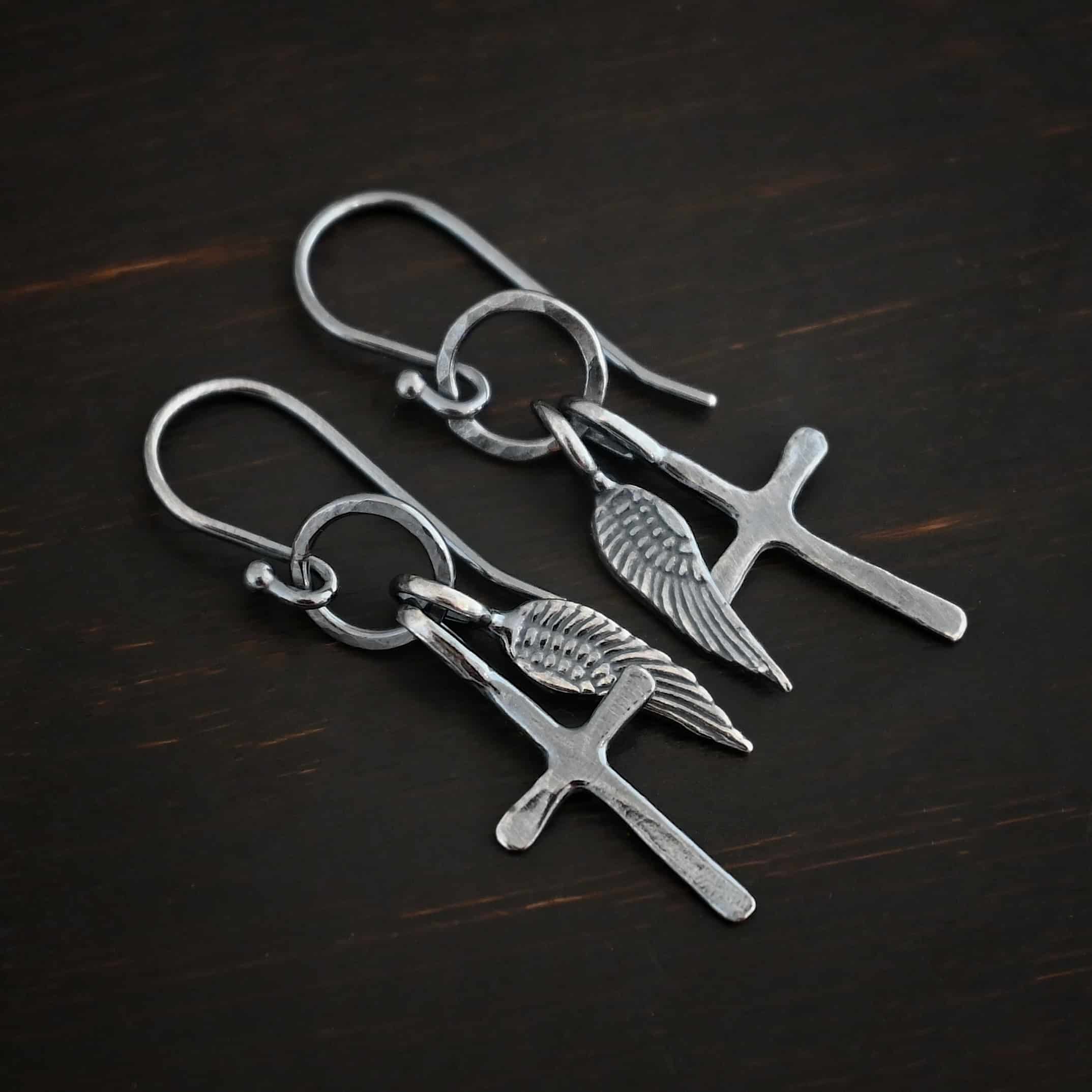 Strength Earrings Photo of the "Strength" Earrings. Made out of oxidized sterling silver. There is a small sized cross that is flat and smooth. It has a loop soldered to the top of it. There is a small sized angel wing pendant next to it, also with a soldered loop on the top. Both of them hang down from a hammered and oxidized circle that hangs on a french hook ear wire. Earrings are lying against a dark background.