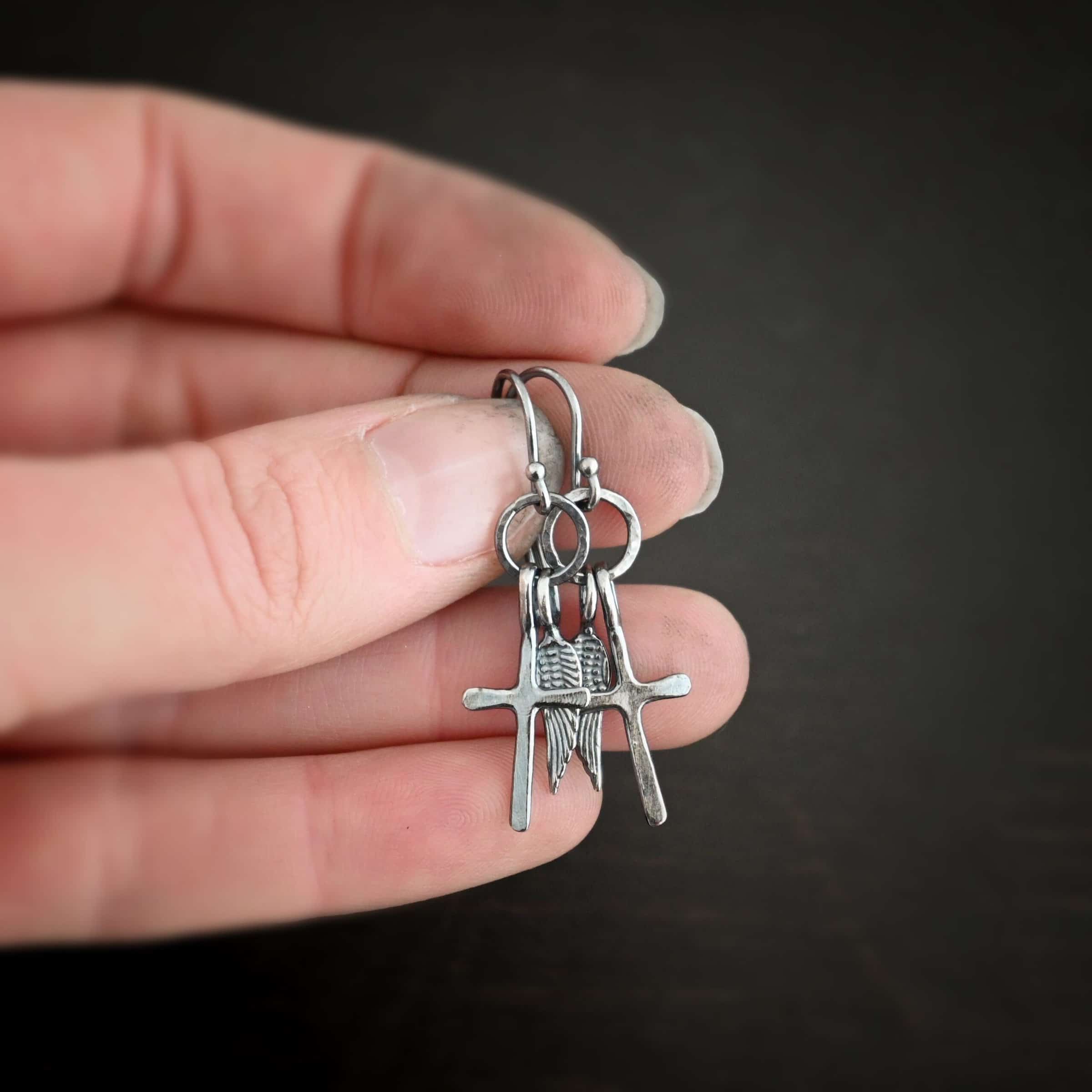 Strength Earrings Photo of the "Strength" Earrings. Made out of oxidized sterling silver. There is a small sized cross that is flat and smooth. It has a loop soldered to the top of it. There is a small sized angel wing pendant next to it, also with a soldered loop on the top. Both of them hang down from a hammered and oxidized circle that hangs on a french hook ear wire. Earrings are held in the fingertips of a hand to demonstrate size.