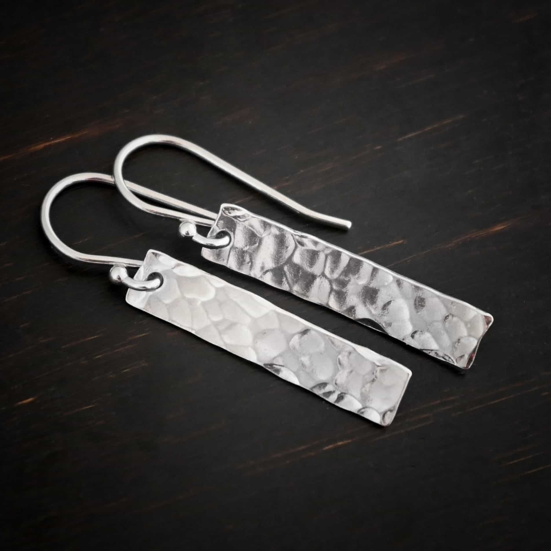 Dive Bar Earrings Photo of the "Dive Bar" Earrings. Made out of sterling silver. Earrings have french hook ear wires. Attached to the bottom of the ear wires is a rectangle piece of sterling silver that is hammered and shiny. Earrings are lying against a dark background.