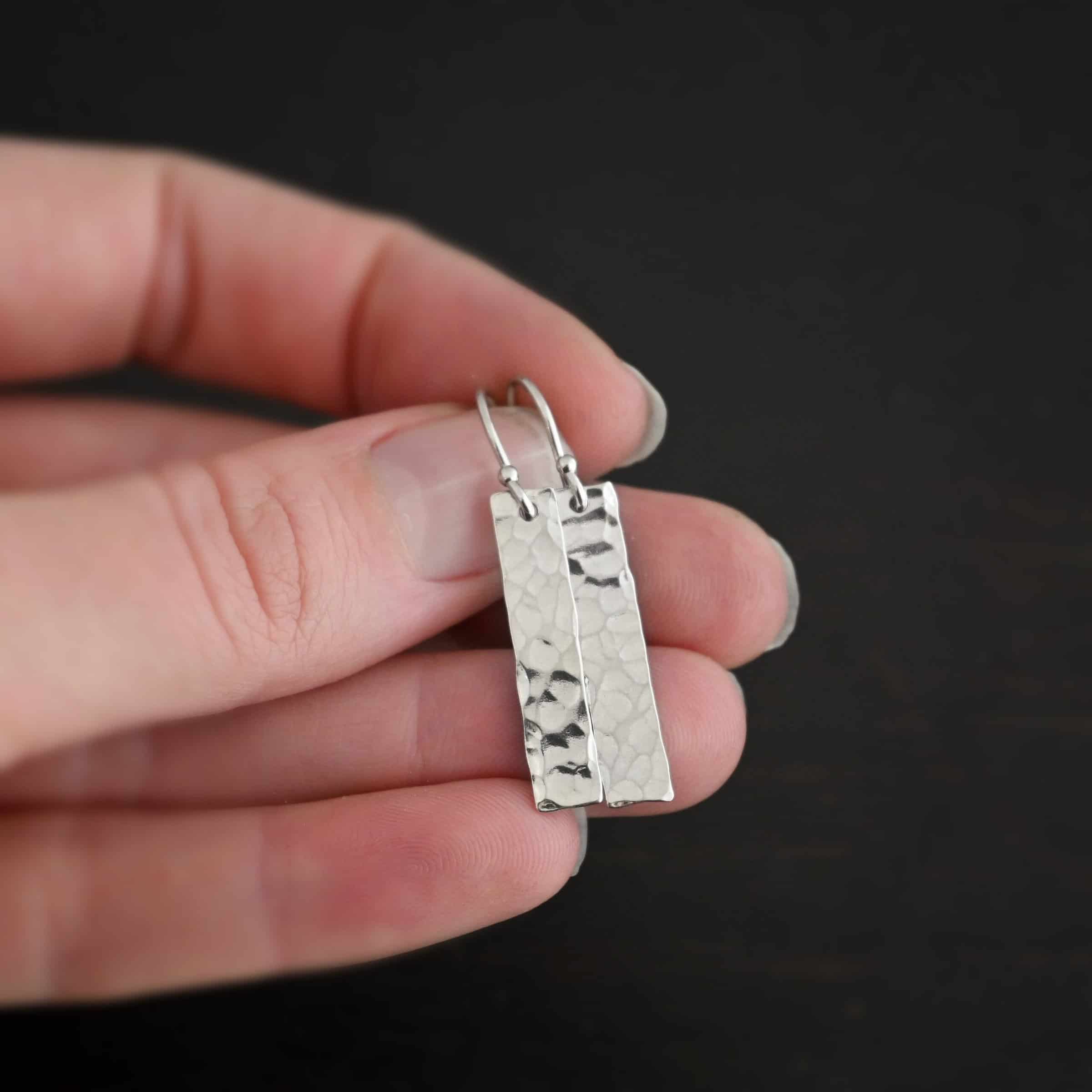 Dive Bar Earrings Photo of the "Dive Bar" Earrings. Made out of sterling silver. Earrings have french hook ear wires. Attached to the bottom of the ear wires is a rectangle piece of sterling silver that is hammered and shiny. Earrings are held in the fingertips of a hand.
