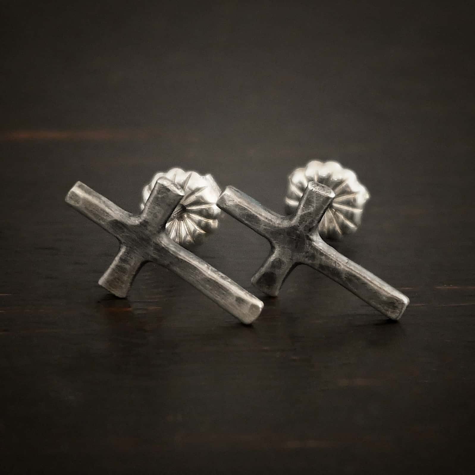 Healer Earrings Photo of the "Healer" earrings. The small crosses are hammered and oxidized, with posts on the back of them and silver earring backs. Earrings are lying against a dark background.