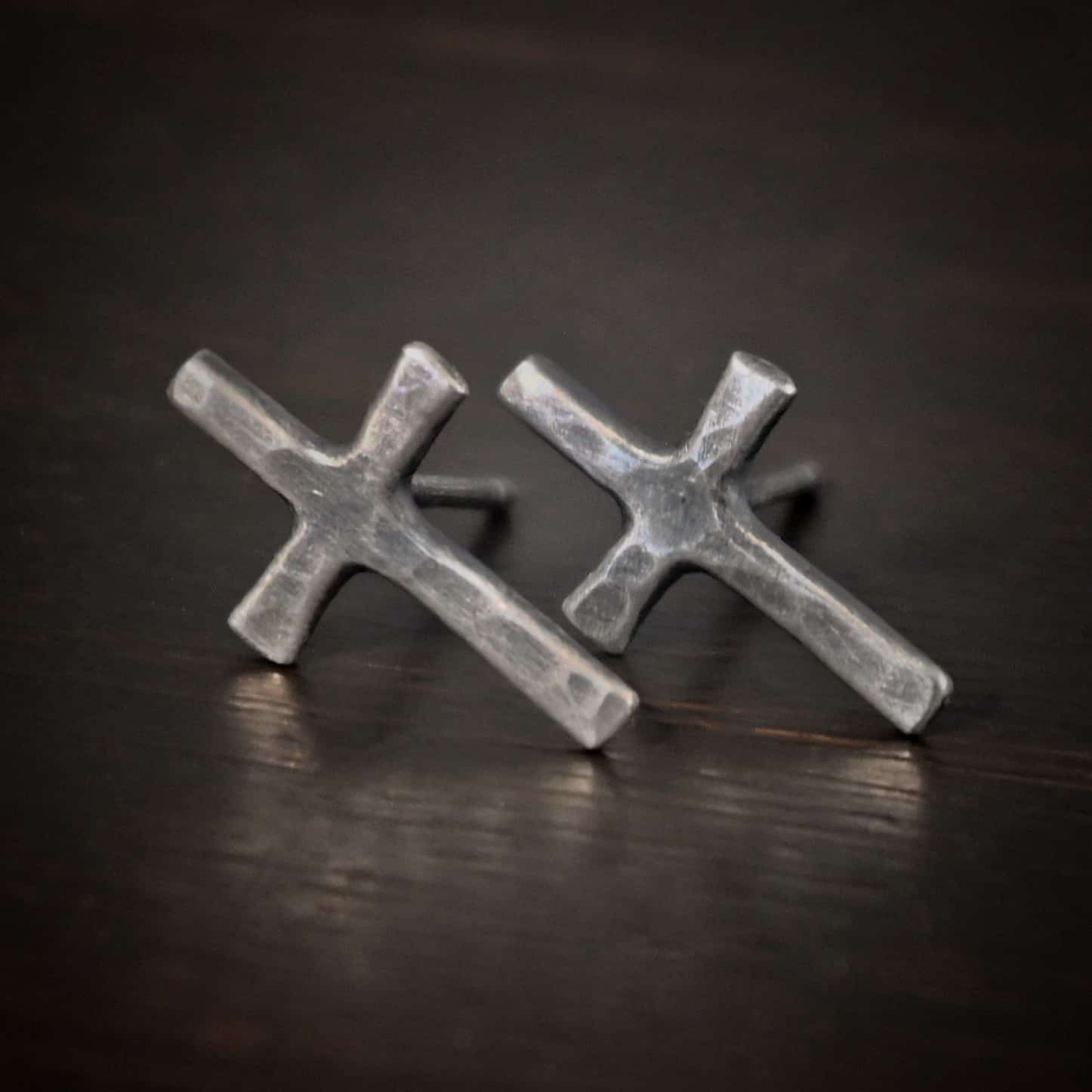 Healer Earrings Photo of the "Healer" earrings. The small crosses are hammered and oxidized, with posts on the back of them. Earrings are lying against a dark background.
