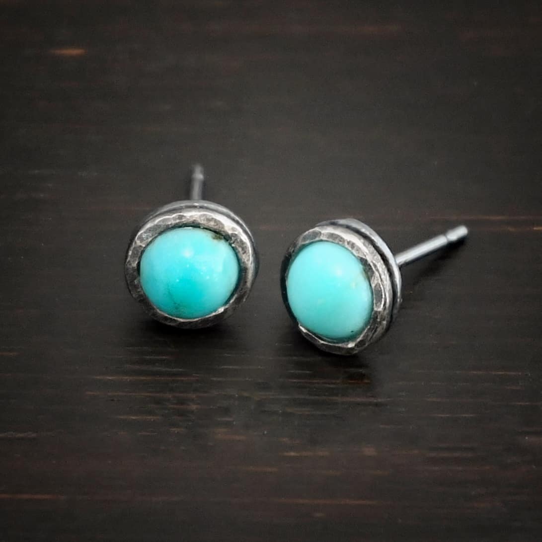 Photo of the "Amelia Earhart" Earrings. Made out of sterling silver and turquoise. Earrings are simple post earrings. Each earring has a small circle turquoise that is set in silver that is hammered and oxidized. Earrings are lying against a dark background.