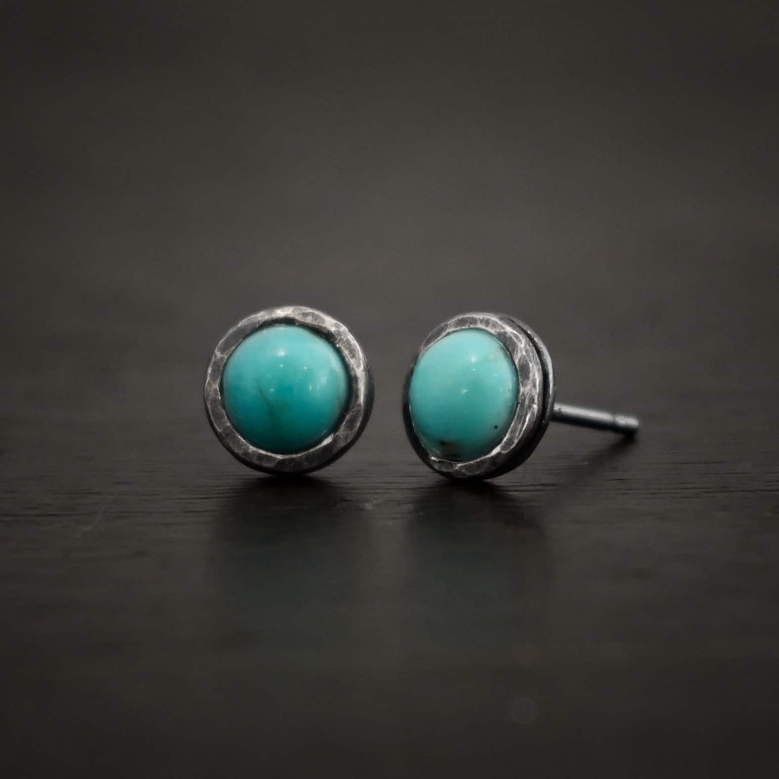 Photo of the "Amelia Earhart" Earrings. Made out of sterling silver and turquoise. Earrings are simple post earrings. Each earring has a small circle turquoise that is set in silver that is hammered and oxidized. Earrings are lying against a dark background.