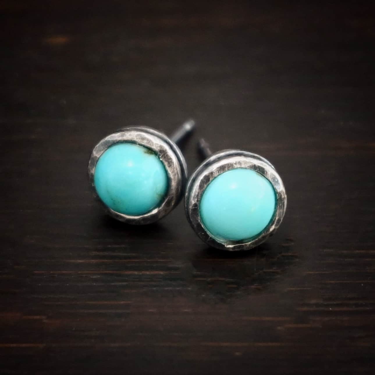 Photo of the "Amelia Earhart" Earrings. Made out of sterling silver and turquoise. Earrings are simple post earrings. Each earring has a small circle turquoise that is set in silver that is hammered and oxidized. Earrings are lying against a dark background.