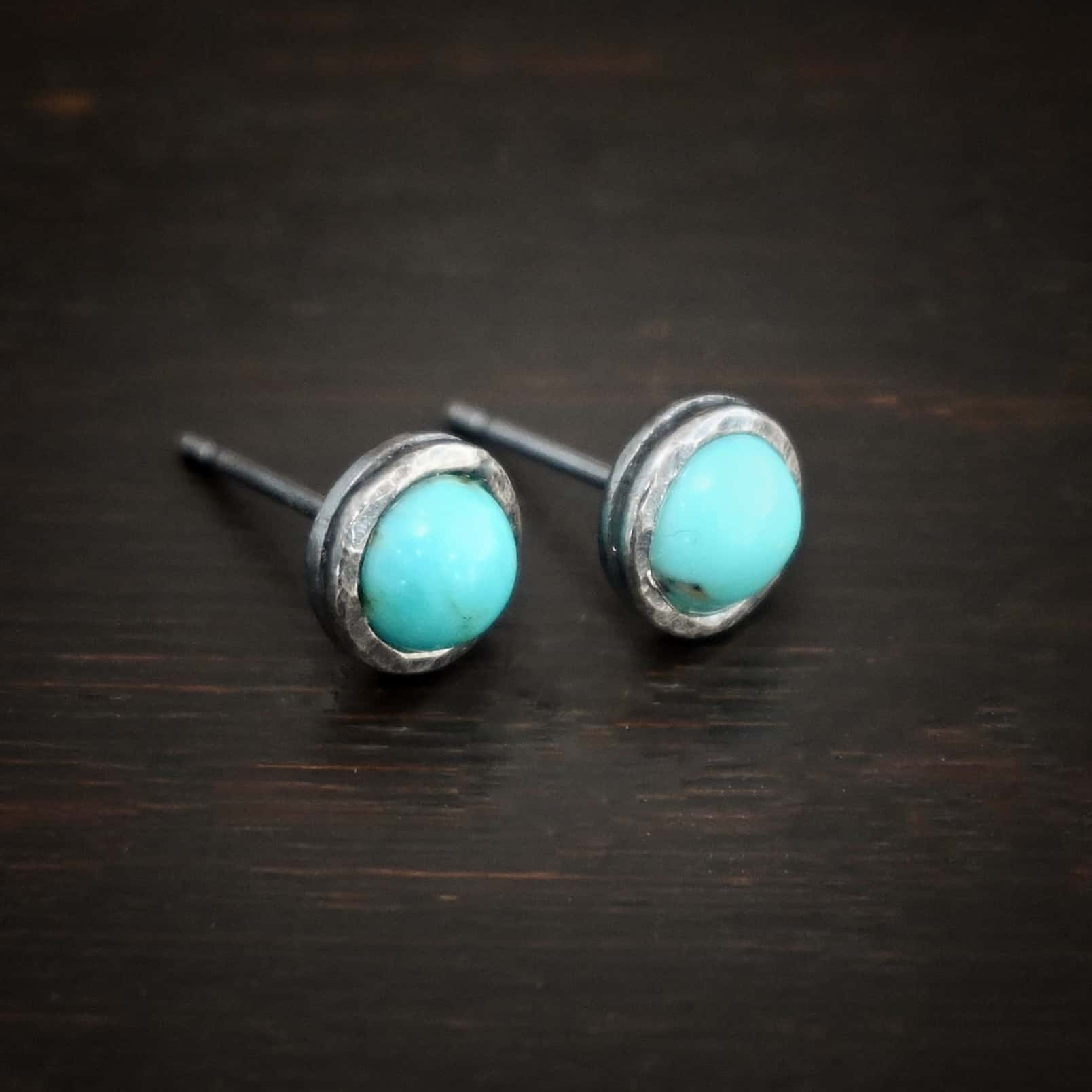 Photo of the "Amelia Earhart" Earrings. Made out of sterling silver and turquoise. Earrings are simple post earrings. Each earring has a small circle turquoise that is set in silver that is hammered and oxidized. Earrings are lying against a dark background.