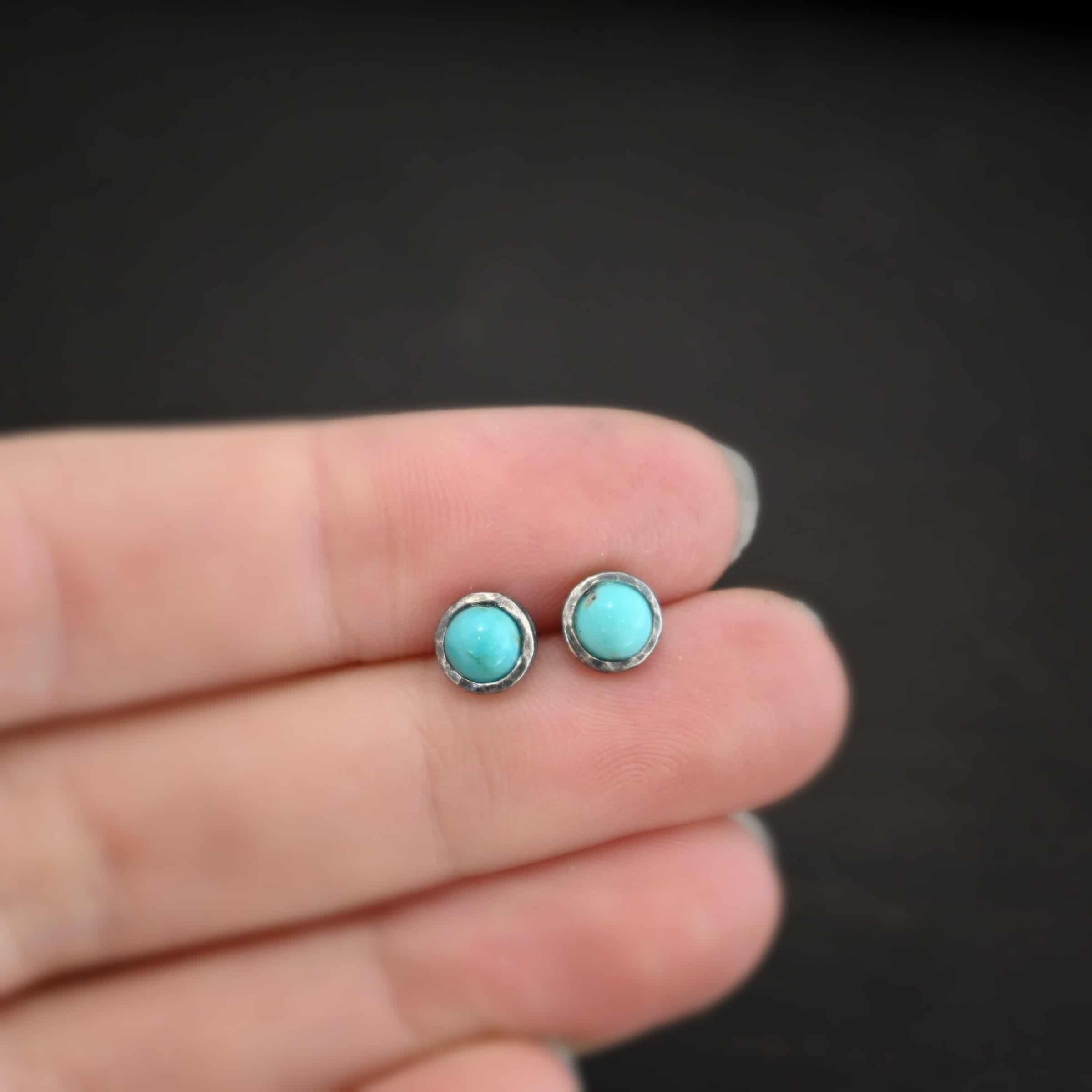 Photo of the "Amelia Earhart" Earrings. Made out of sterling silver and turquoise. Earrings are simple post earrings. Each earring has a small circle turquoise that is set in silver that is hammered and oxidized. Earrings are held in the fingertips of a hand.