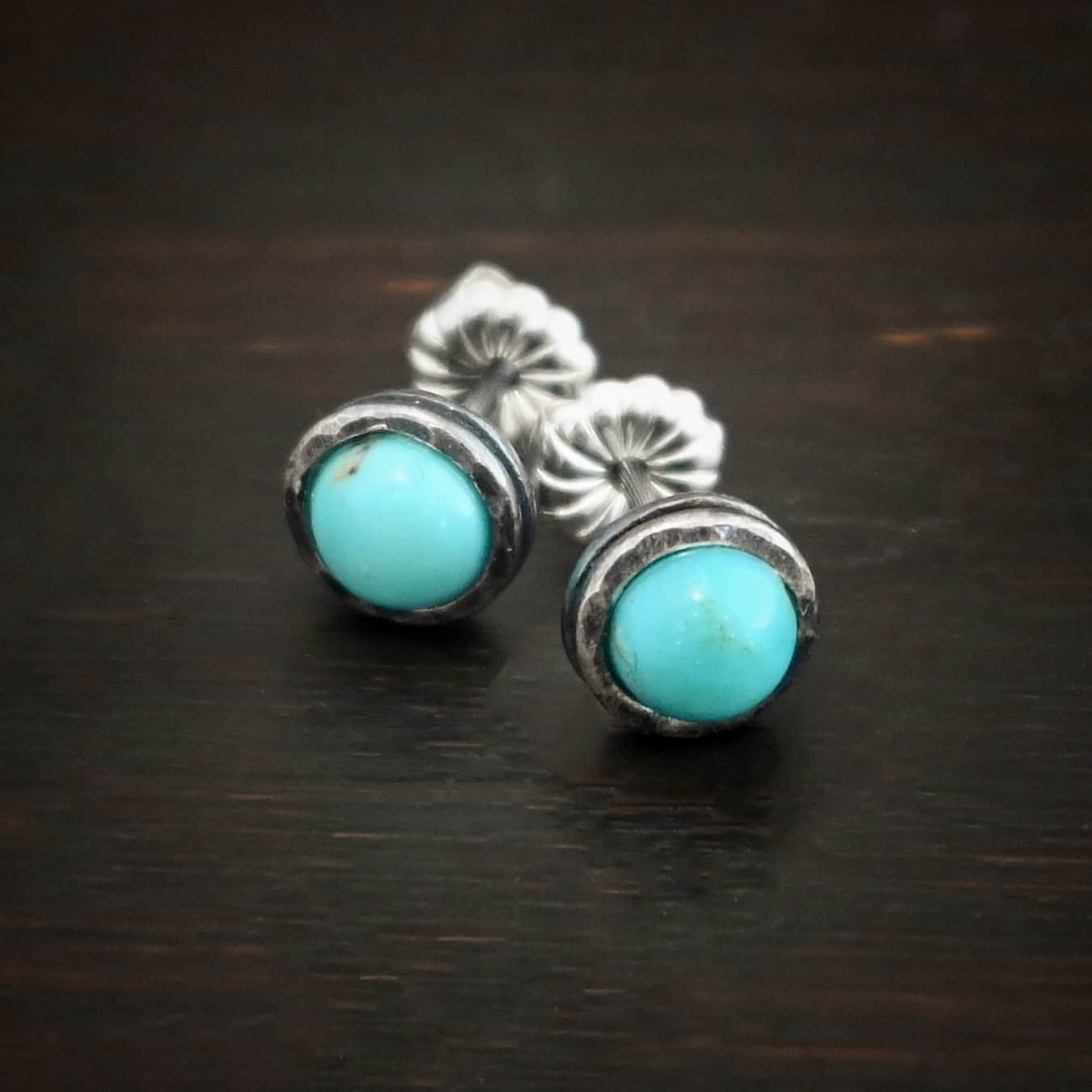 Photo of the "Amelia Earhart" Earrings. Made out of sterling silver and turquoise. Earrings are simple post earrings. Each earring has a small circle turquoise that is set in silver that is hammered and oxidized. Earrings are lying against a dark background.