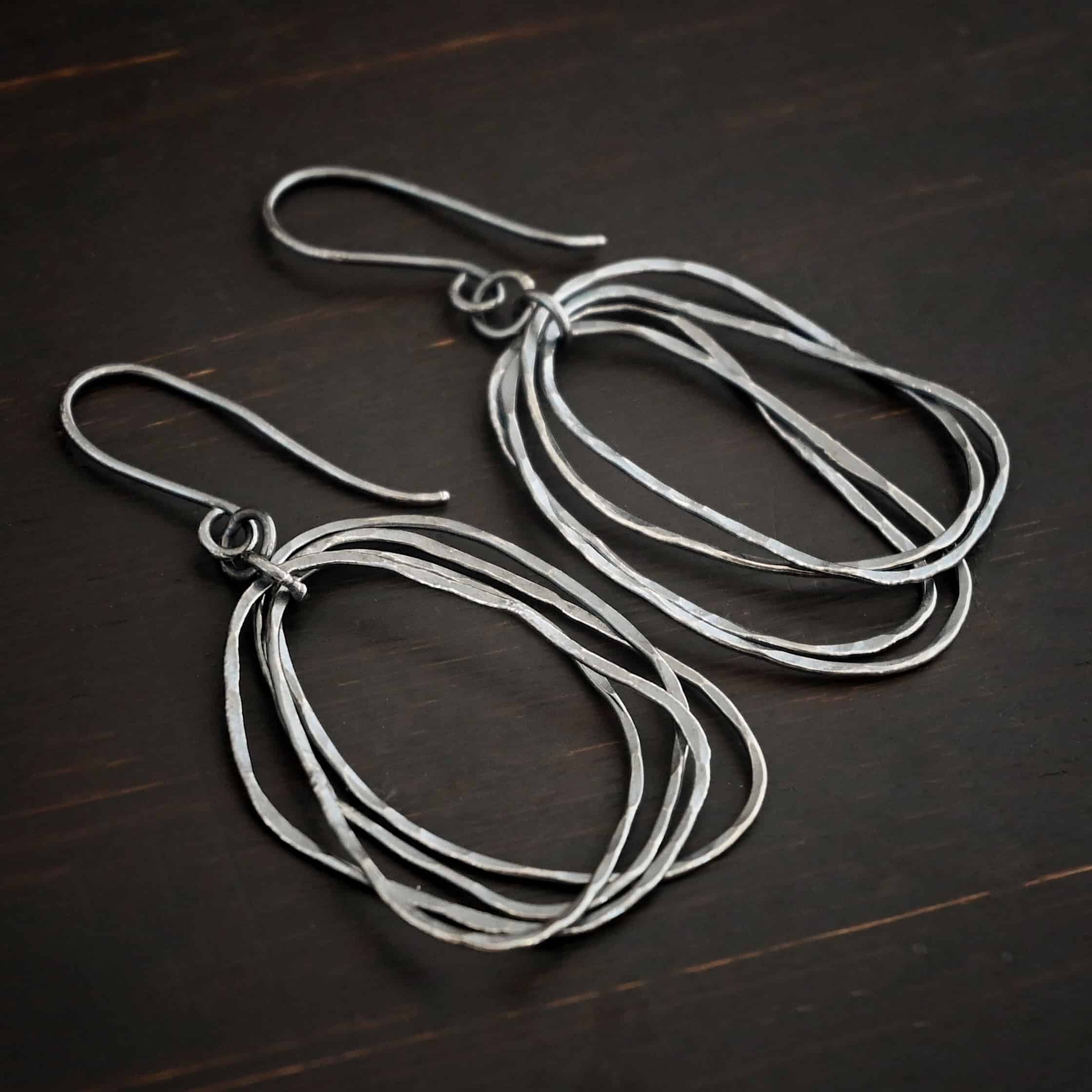 Tangled Earrings Photo of the "Tangled" Earrings. Made out of sterling silver and oxidized. Earrings have french hook ear wires with jump rings attached to the bottom. There is another jump ring attached to the first jump ring. Going through the center of this jump ring are individual circles of sterling silver that are oxidized and hammered. The circles are unevenly hammered, and are not in line with each other, so you can see each circle. Earrings are lying against a dark background.