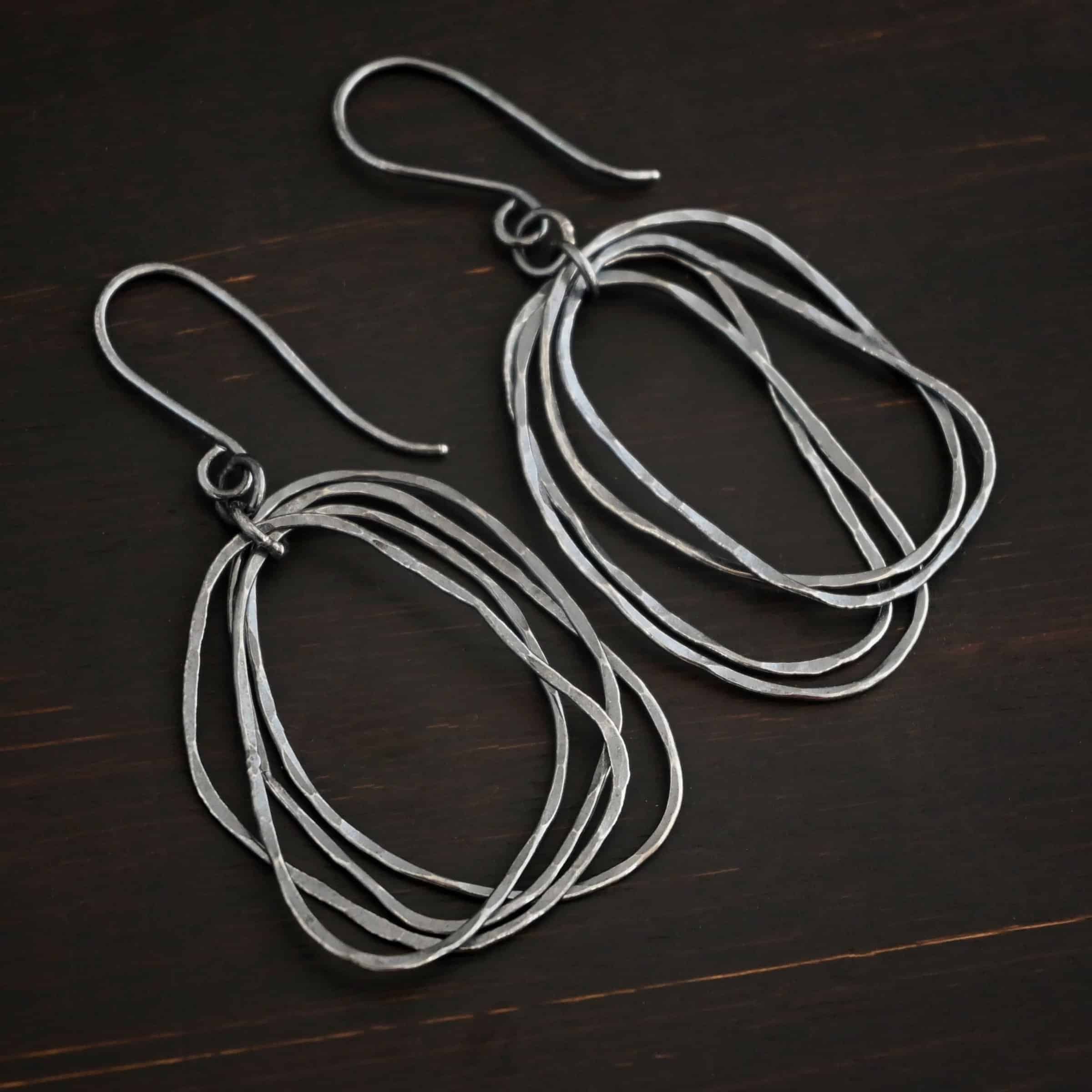 Tangled Earrings Photo of the "Tangled" Earrings. Made out of sterling silver and oxidized. Earrings have french hook ear wires with jump rings attached to the bottom. There is another jump ring attached to the first jump ring. Going through the center of this jump ring are individual circles of sterling silver that are oxidized and hammered. The circles are unevenly hammered, and are not in line with each other, so you can see each circle. Earrings are lying against a dark background.