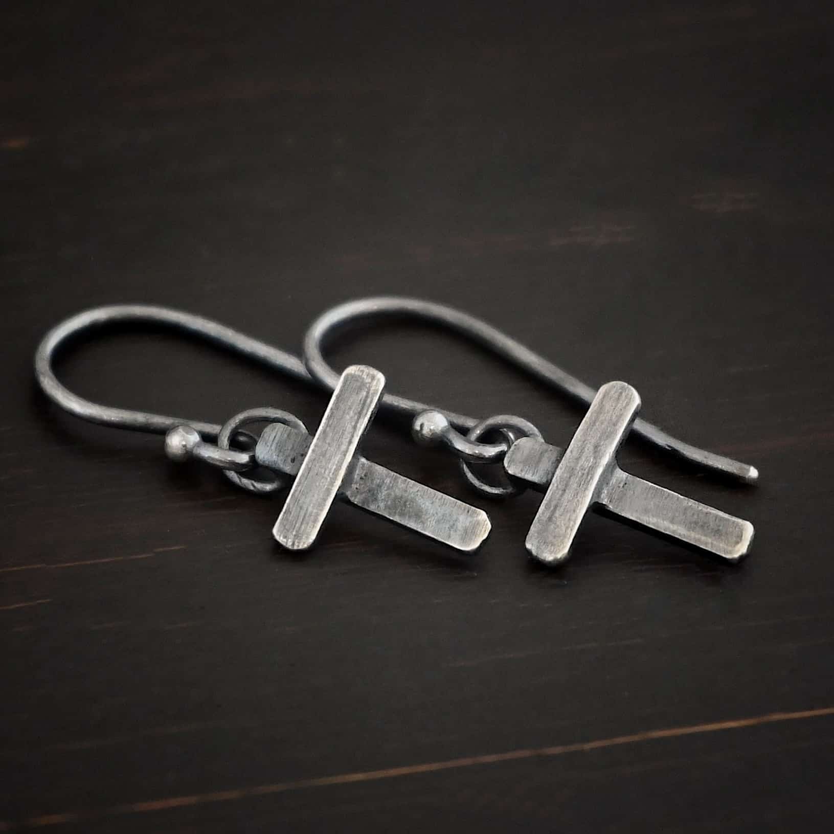 Photo of the "Old Rugged Cross" Earrings. Made out of sterling silver and oxidized. Earrings have a french hook ear wire with a jump ring on the bottom of it. Soldered to the jump ring is a small handmade cross charm. The cross has a small rectangle piece on the back set vertically, and a shorter rectangle piece soldered on top to the back piece. The shorter piece is set horizontally across the front of the back piece.