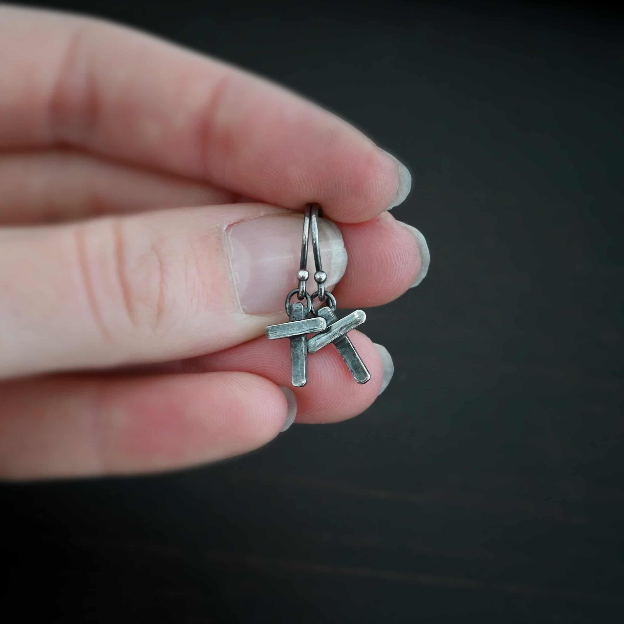 Photo of the "Old Rugged Cross" Earrings. Made out of sterling silver and oxidized. Earrings have a french hook ear wire with a jump ring on the bottom of it. Soldered to the jump ring is a small handmade cross charm. The cross has a small rectangle piece on the back set vertically, and a shorter rectangle piece soldered on top to the back piece. The shorter piece is set horizontally across the front of the back piece. Earrings are held in the fingertips of a hand.