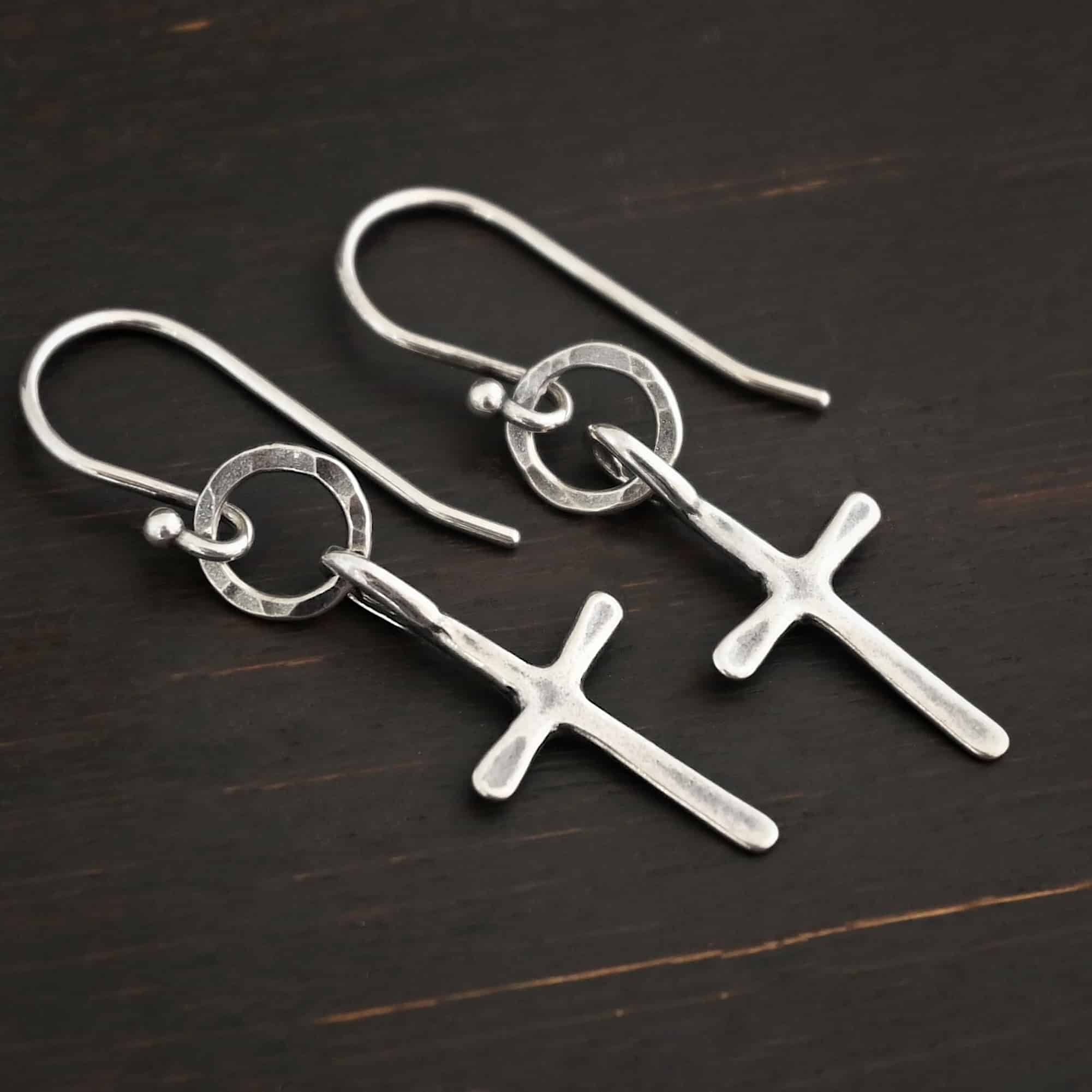 Photo of the "Redeemer" Earrings. Made out of sterling silver. Earrings have a very small cross that is hammered flat, but smooth, with a loop soldered to the top of it. It hangs from a small, hammered, circle that is shiny. The circle hangs from a french hook ear wire that is oxidized. Earring is lying against a dark background.