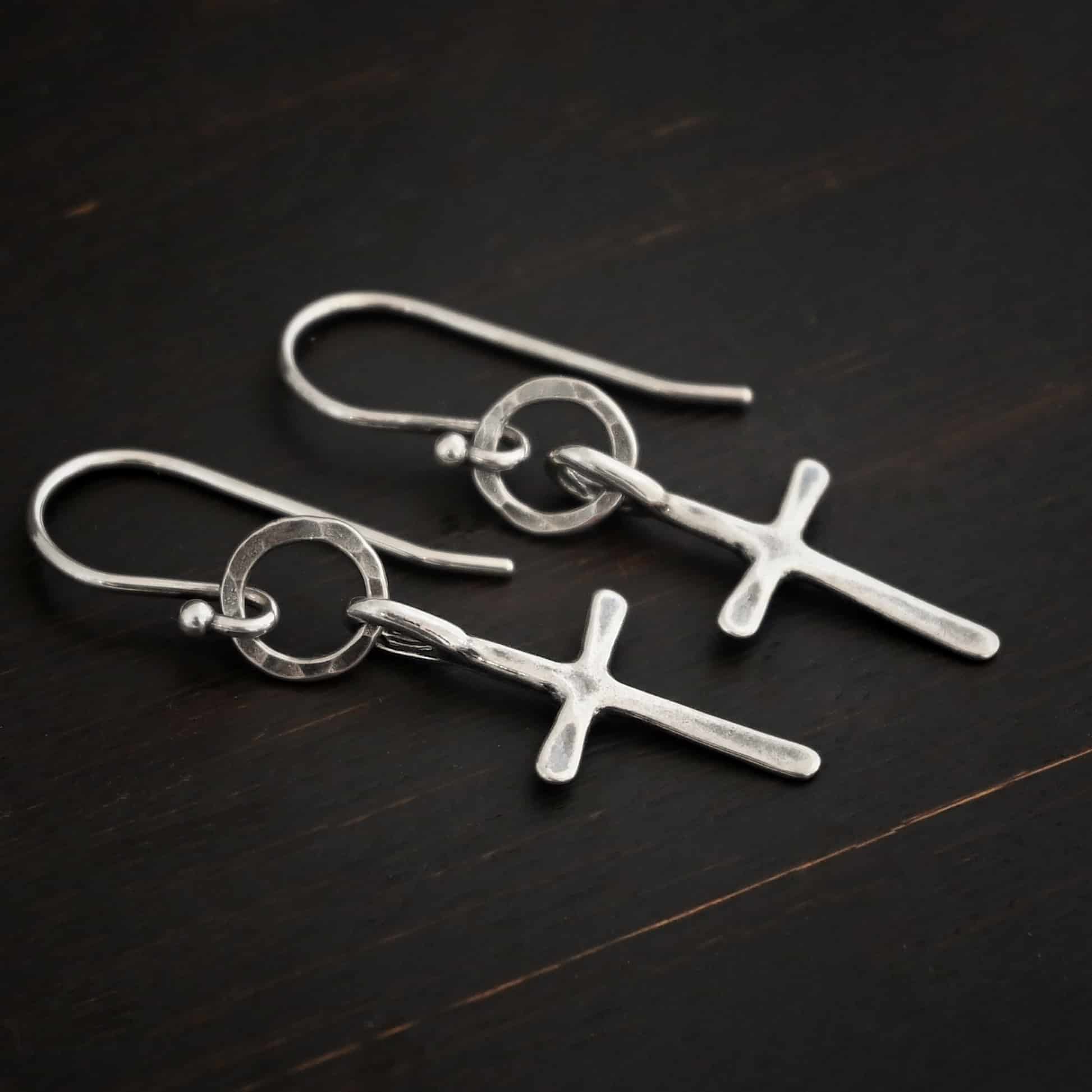 Photo of the "Redeemer" Earrings. Made out of sterling silver. Earrings have a very small cross that is hammered flat, but smooth, with a loop soldered to the top of it. It hangs from a small, hammered, circle that is oxidized. The circle hangs from a french hook ear wire that is oxidized. Earring is lying against a dark background.