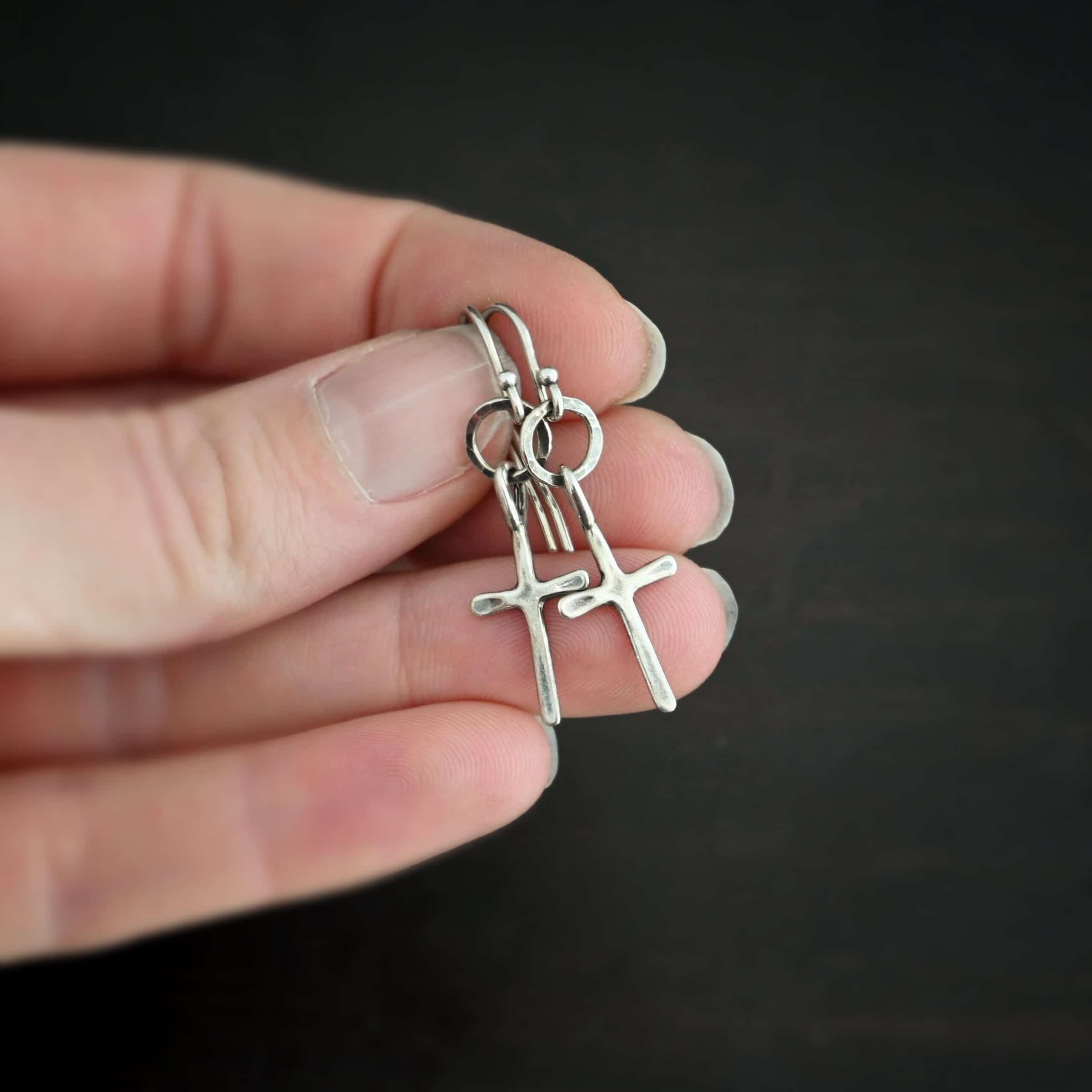 Photo of the "Redeemer" Earrings. Made out of sterling silver. Earrings have a very small cross that is hammered flat, but smooth, with a loop soldered to the top of it. It hangs from a small, hammered, circle that is oxidized. The circle hangs from a french hook ear wire that is oxidized. Earrings are held in the fingertips of a hand.