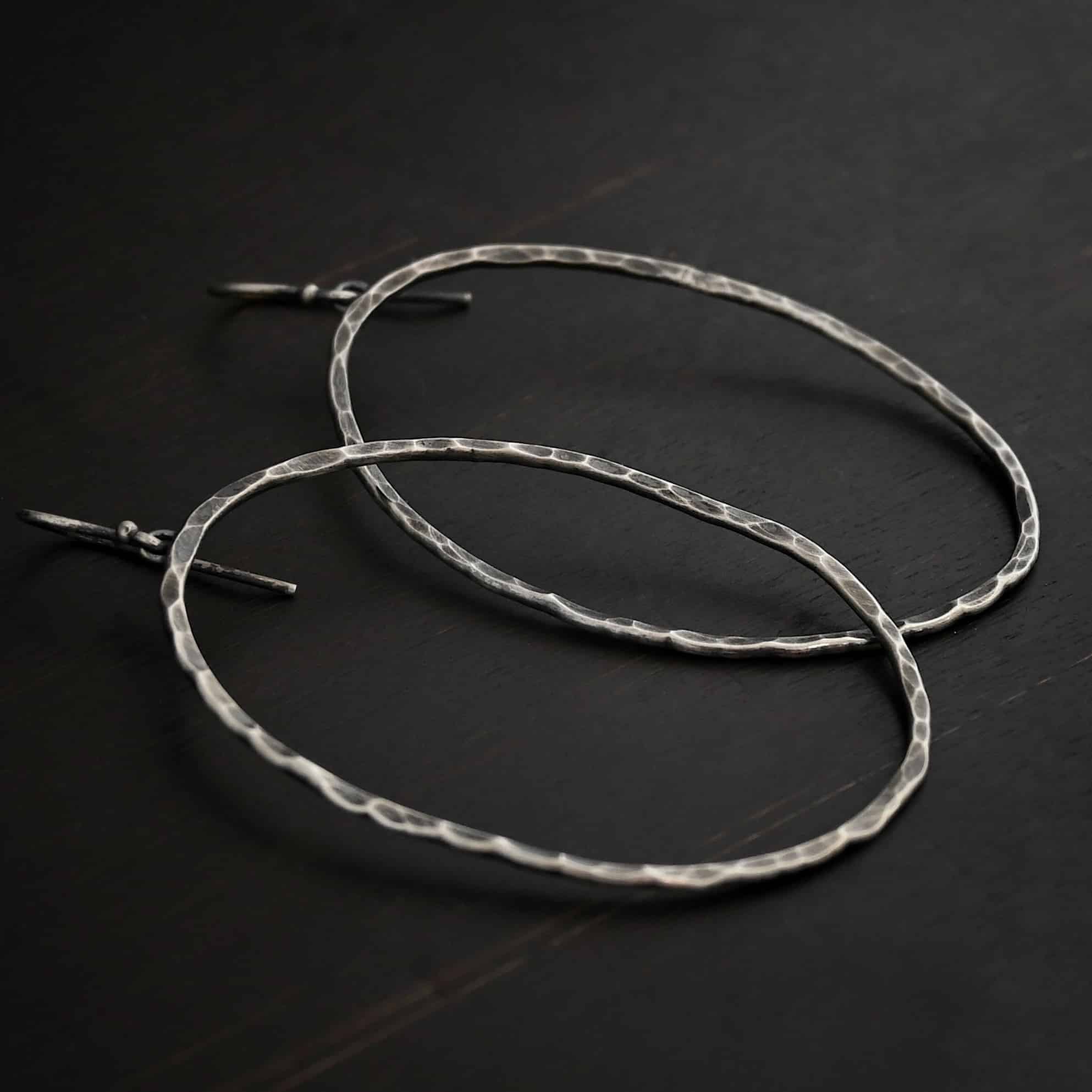Photo of the "Haines Beach" Hoop Earrings. Made out of sterling silver and oxidized. Earrings have french hook ear wires. Attached to the bottom of the ear wires are small loops. The small loops are soldered to the back top of large hoop earrings that are hammered and oxidized ovals. Earrings are lying against a dark background.