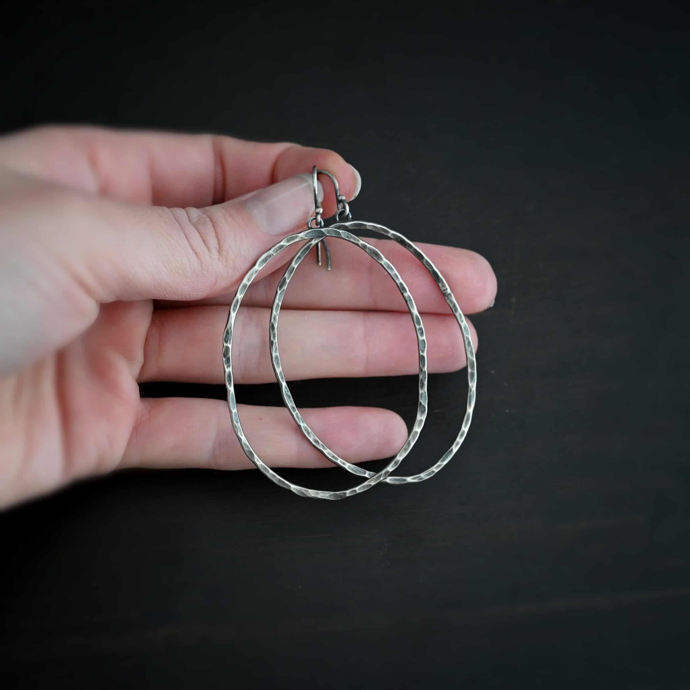 Photo of the "Haines Beach" Hoop Earrings. Made out of sterling silver and oxidized. Earrings have french hook ear wires. Attached to the bottom of the ear wires are small loops. The small loops are soldered to the back top of large hoop earrings that are hammered and oxidized ovals. Earrings are held in the fingertips of a hand.