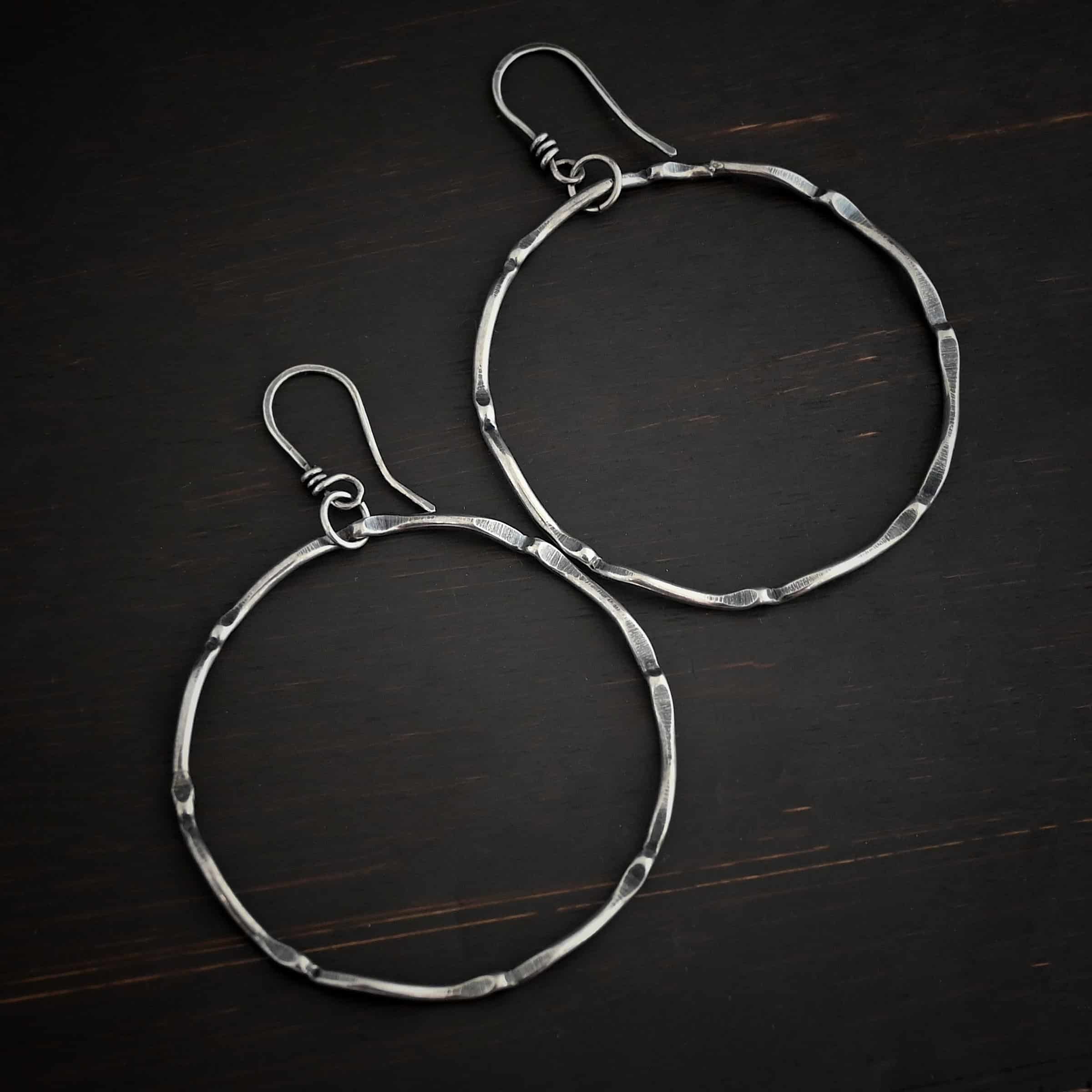 Photo of the "Tidal Wave" Hoop Earrings. Made out of sterling silver and oxidized. Earrings have french hook ear wires with a loop on the bottom. Going through the bottom loop is a jump ring. Going through the center of the jump ring is a large circle hoop that is oxidized and bent in places all around the circle to give it a textured loop. Earrings are lying against a dark background.
