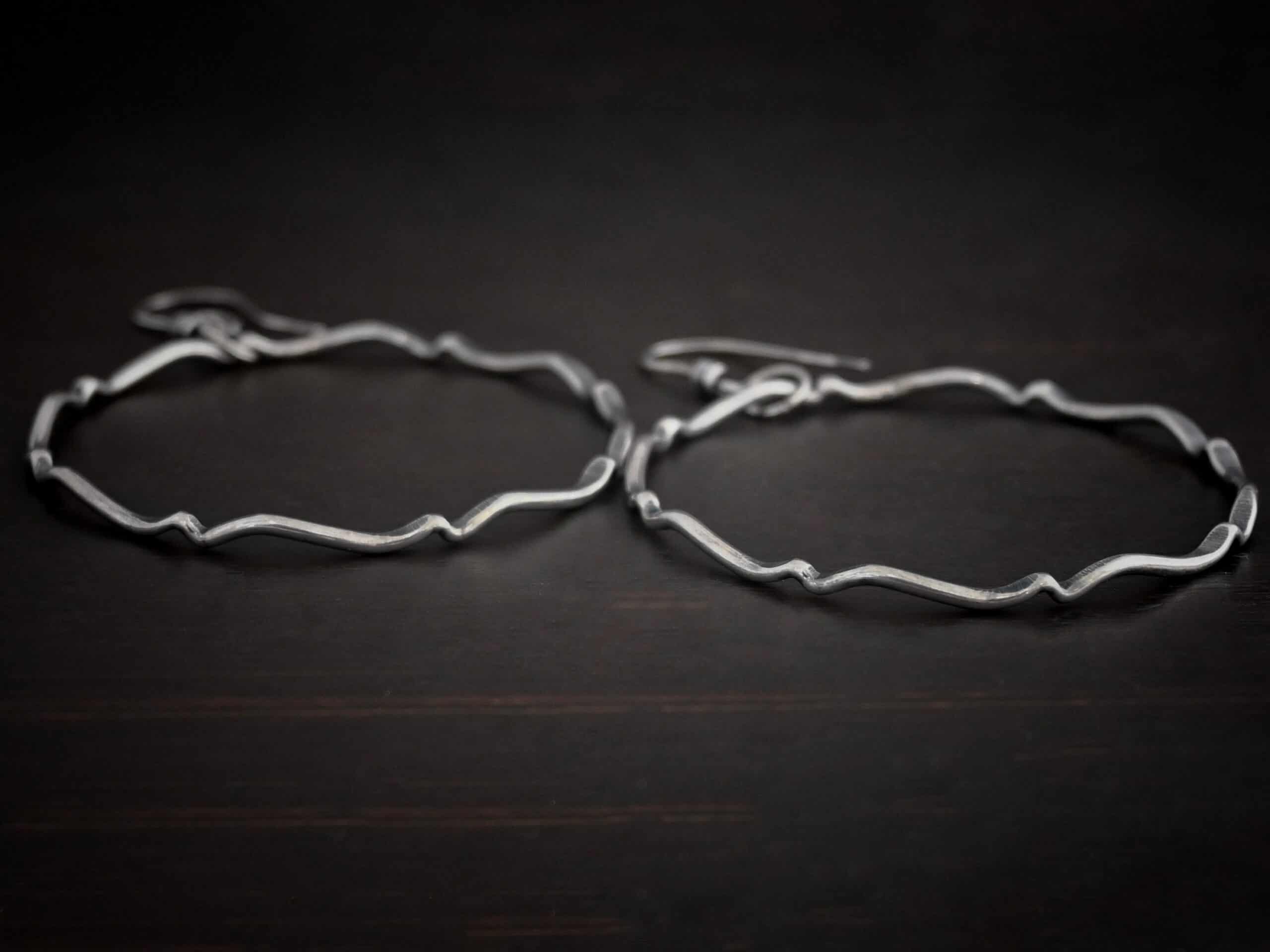 Photo of the "Tidal Wave" Hoop Earrings. Made out of sterling silver and oxidized. Earrings have french hook ear wires with a loop on the bottom. Going through the bottom loop is a jump ring. Going through the center of the jump ring is a large circle hoop that is oxidized and bent in places all around the circle to give it a textured loop. Earrings are lying against a dark background.