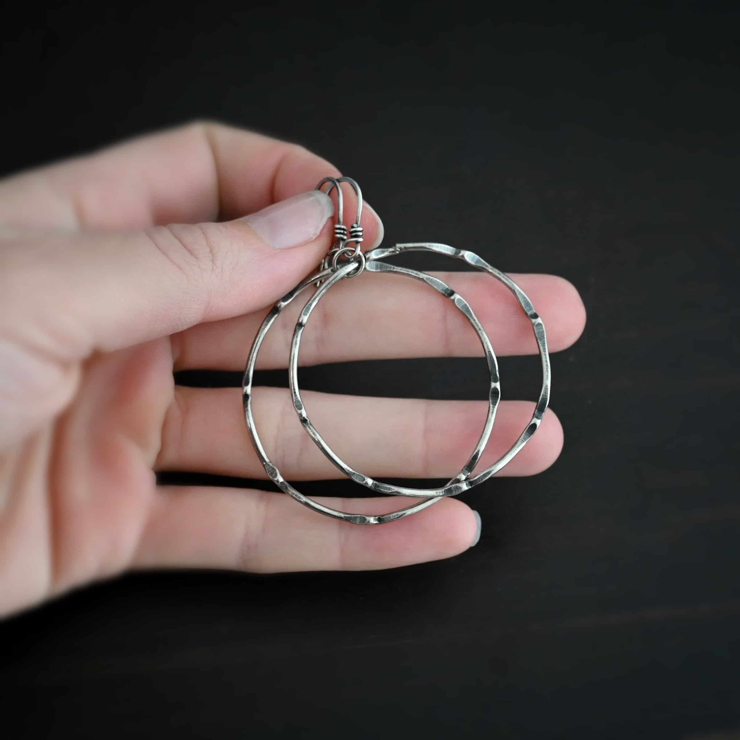 Photo of the "Tidal Wave" Hoop Earrings. Made out of sterling silver and oxidized. Earrings have french hook ear wires with a loop on the bottom. Going through the bottom loop is a jump ring. Going through the center of the jump ring is a large circle hoop that is oxidized and bent in places all around the circle to give it a textured loop. Earrings are held in the fingertips of a hand.
