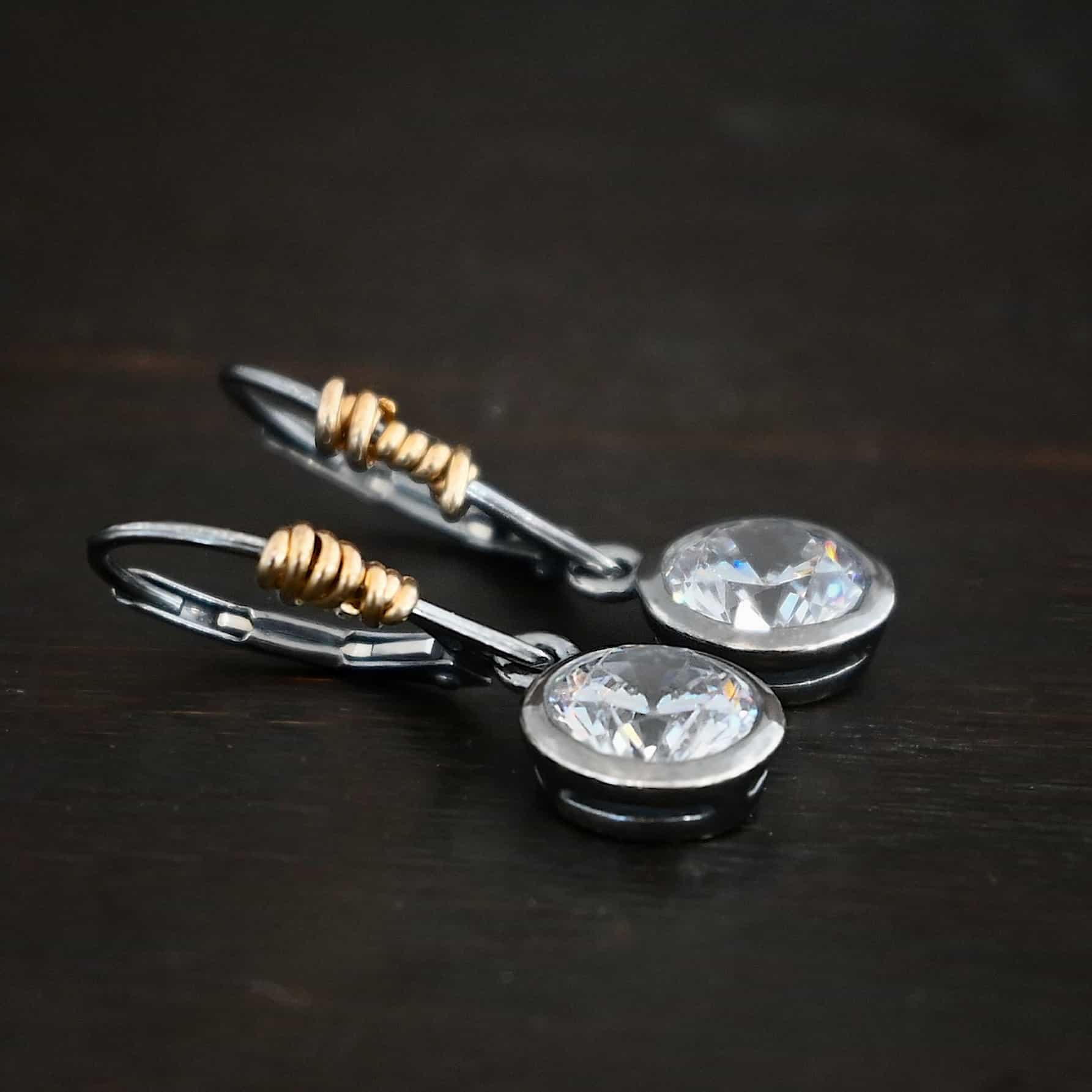 Photo of the "Audrey Hepburn" Earrings. Made out of sterling silver, gold fill, and moissanites. Earrings have sterling silver, oxidized, leverback ear wires with gold filled wrap around the bottom part of the ear wires on the left side. Attached to the bottom loop is a large, circle moissanite that is set in silver. The silver has a loop on the top to connect it to the ear wire. Earrings are lying against a dark background.