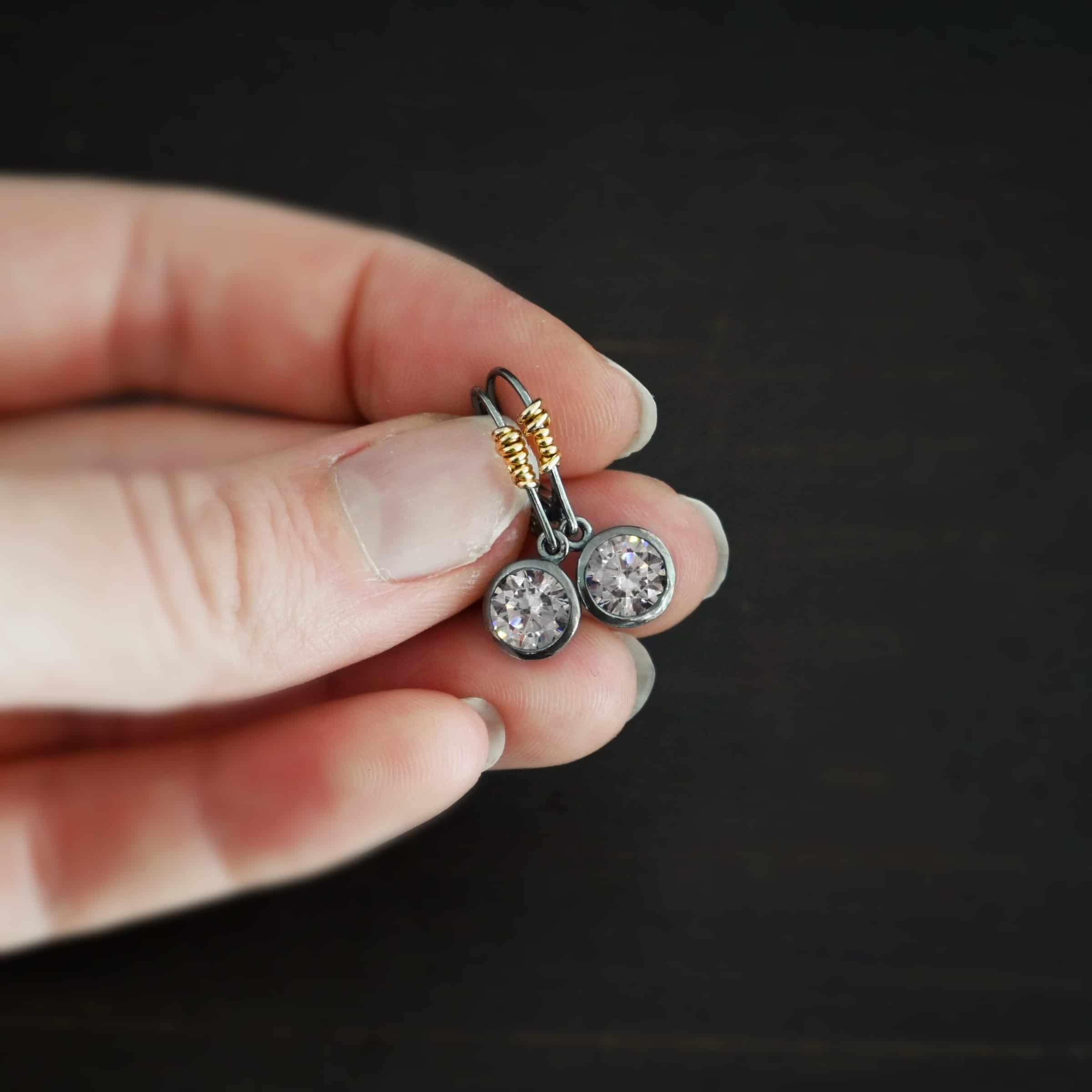 Photo of the "Audrey Hepburn" Earrings. Made out of sterling silver, gold fill, and moissanites. Earrings have sterling silver, oxidized, leverback ear wires with gold filled wrap around the bottom part of the ear wires on the left side. Attached to the bottom loop is a large, circle moissanite that is set in silver. The silver has a loop on the top to connect it to the ear wire. Earrings are held in the fingertips of a hand to demonstrate size.