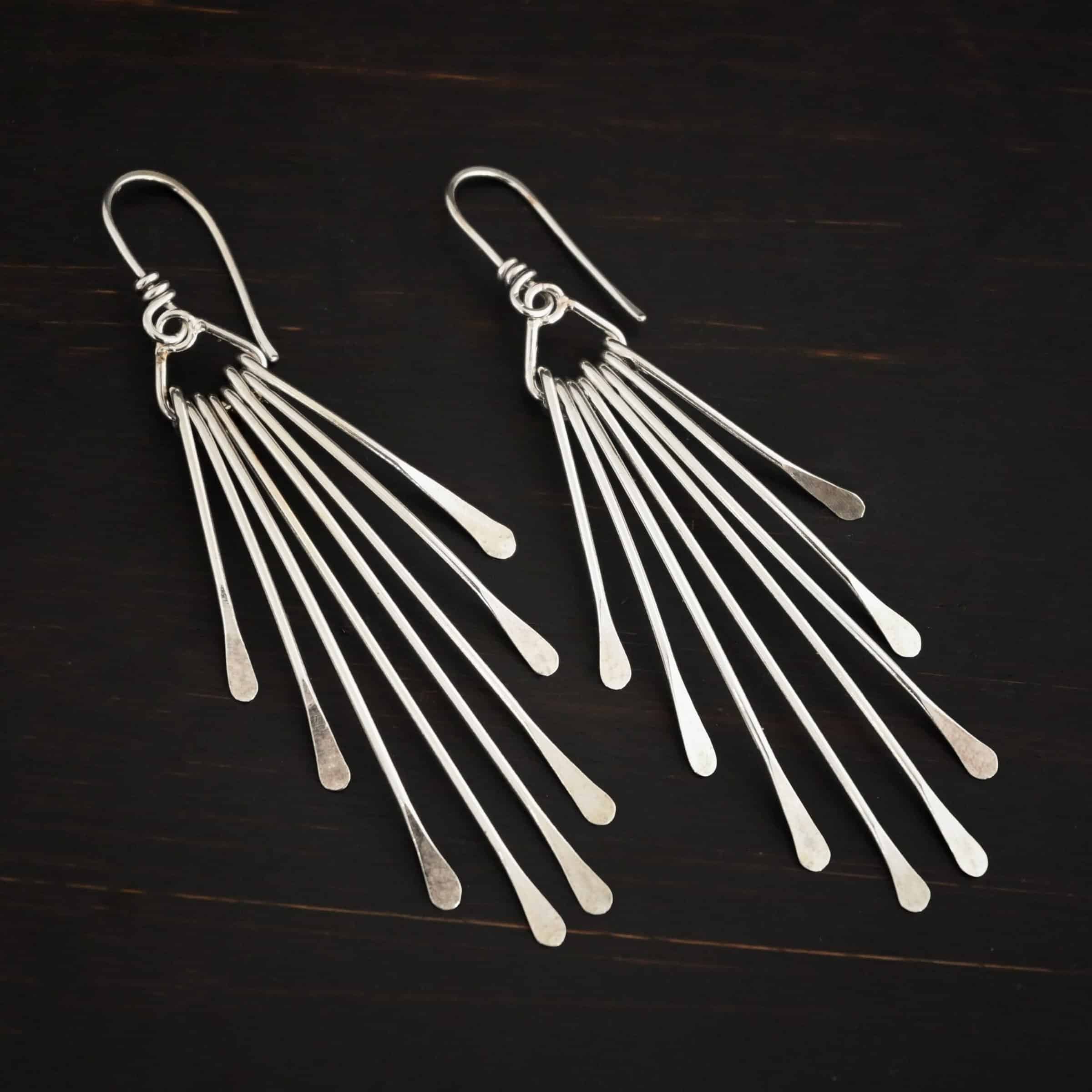 Photo of the "Marilyn Monroe" Earrings. Made out of sterling silver. Earrings have french hook ear wires. Attached to the bottom loop of the ear wires is a teardrop shaped piece of silver. Hanging from this piece are eight separate pieces of silver, all with hammered paddle ends. They all hang straight down. The shortest ones are on the outside, and they get progressively longer until the middle. So they go from short, to long, and back to short. Earrings are lying against a dark background.