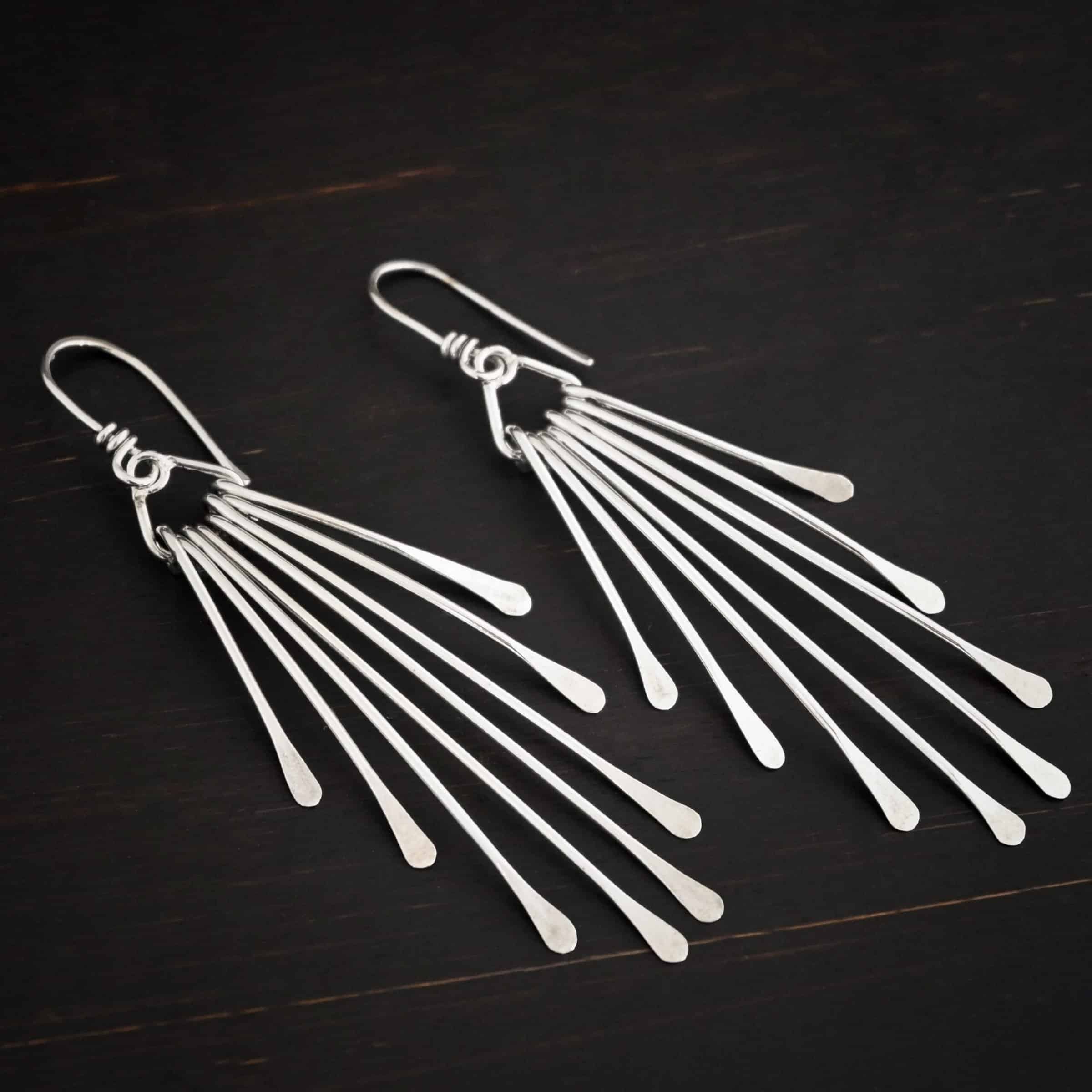 Photo of the "Marilyn Monroe" Earrings. Made out of sterling silver. Earrings have french hook ear wires. Attached to the bottom loop of the ear wires is a teardrop shaped piece of silver. Hanging from this piece are eight separate pieces of silver, all with hammered paddle ends. They all hang straight down. The shortest ones are on the outside, and they get progressively longer until the middle. So they go from short, to long, and back to short. Earrings are lying against a dark background.