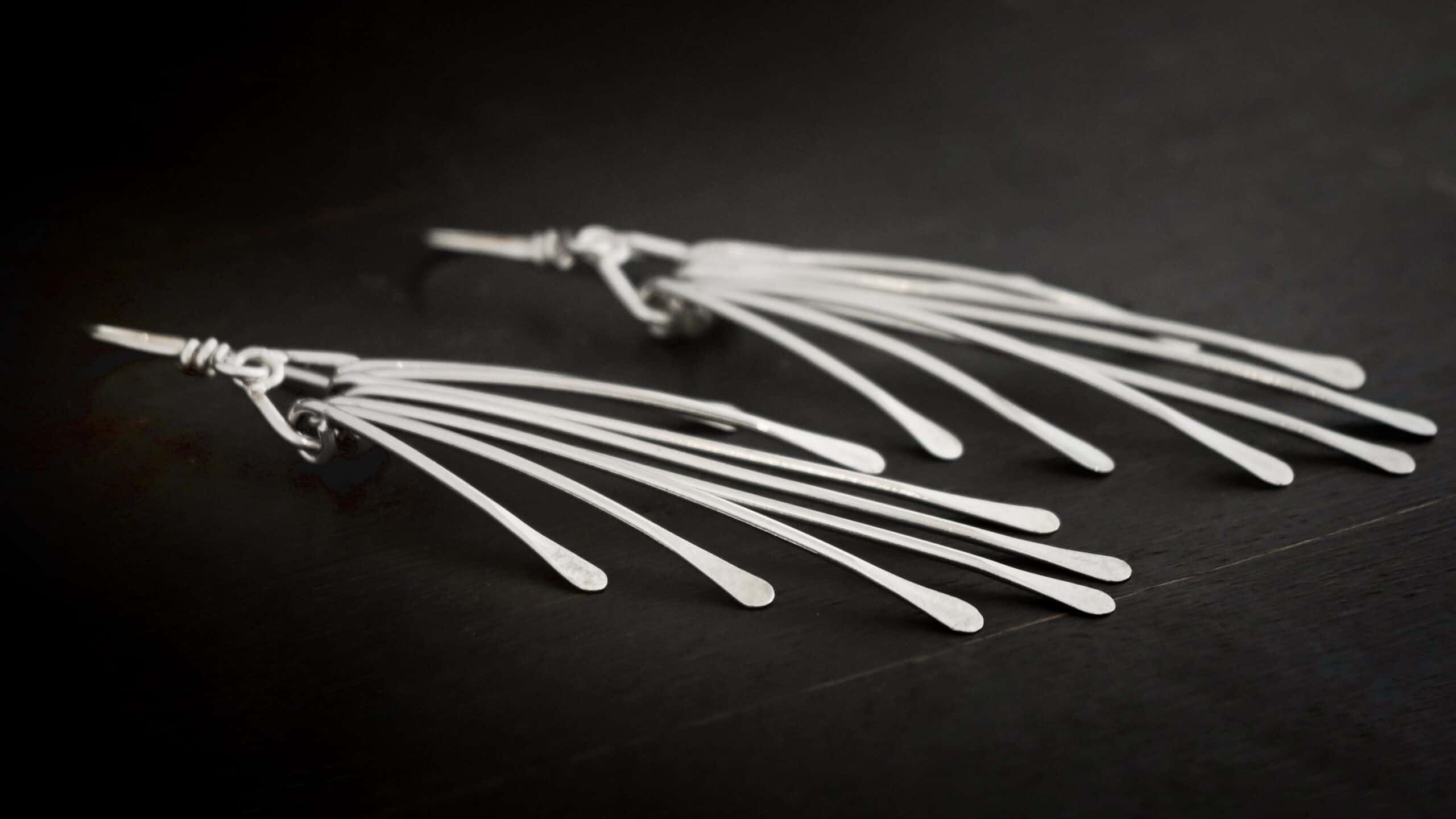 Photo of the "Marilyn Monroe" Earrings. Made out of sterling silver. Earrings have french hook ear wires. Attached to the bottom loop of the ear wires is a teardrop shaped piece of silver. Hanging from this piece are eight separate pieces of silver, all with hammered paddle ends. They all hang straight down. The shortest ones are on the outside, and they get progressively longer until the middle. So they go from short, to long, and back to short. Earrings are lying against a dark background.