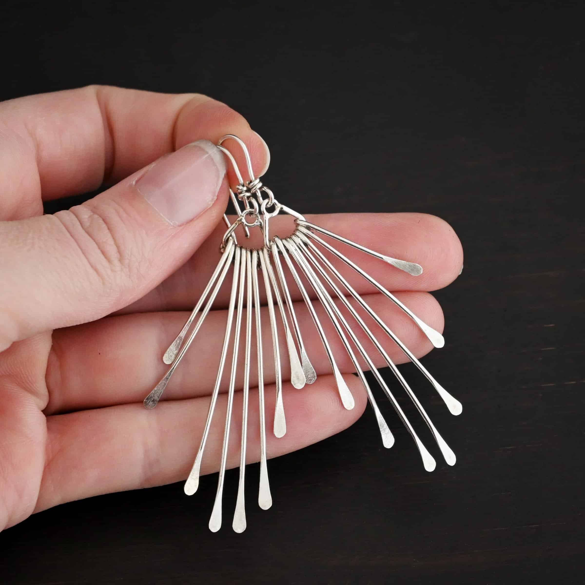 Photo of the "Marilyn Monroe" Earrings. Made out of sterling silver. Earrings have french hook ear wires. Attached to the bottom loop of the ear wires is a teardrop shaped piece of silver. Hanging from this piece are eight separate pieces of silver, all with hammered paddle ends. They all hang straight down. The shortest ones are on the outside, and they get progressively longer until the middle. So they go from short, to long, and back to short. Earrings are held in the fingertips of a hand.