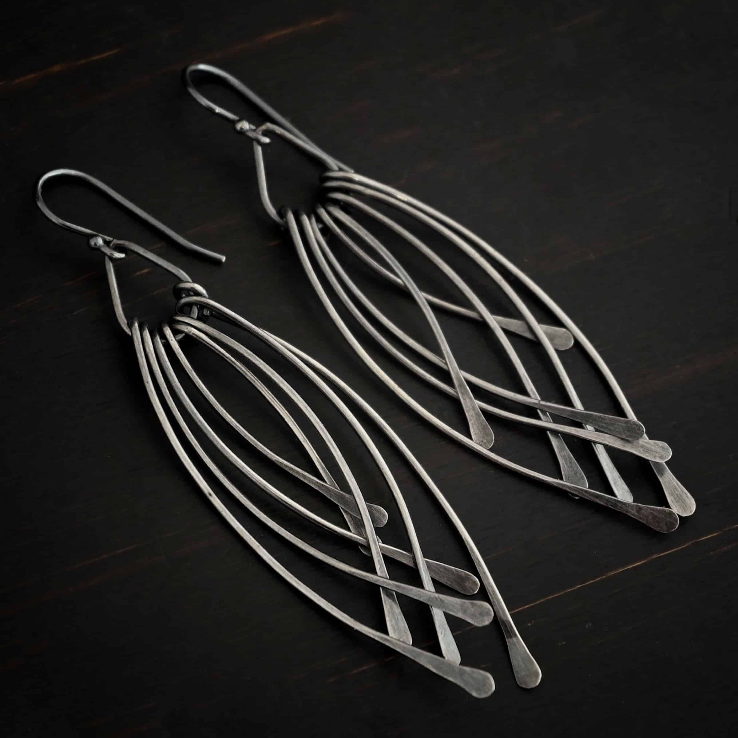Prairie Wind Earrings Photo of the "Prairie Wind" Earrings. Made out of sterling silver and oxidized. Earrings have french hook ear wires. Attached to the bottom of the ear wires is a teardrop shaped loop of silver. Attached to the bottom of the teardrop loop are eight pieces of silver that all have hammered paddle ends. Four pieces of silver curve towards the right, and the other four pieces curve towards the left, so they overlap each other. Earrings are lying against a dark background.
