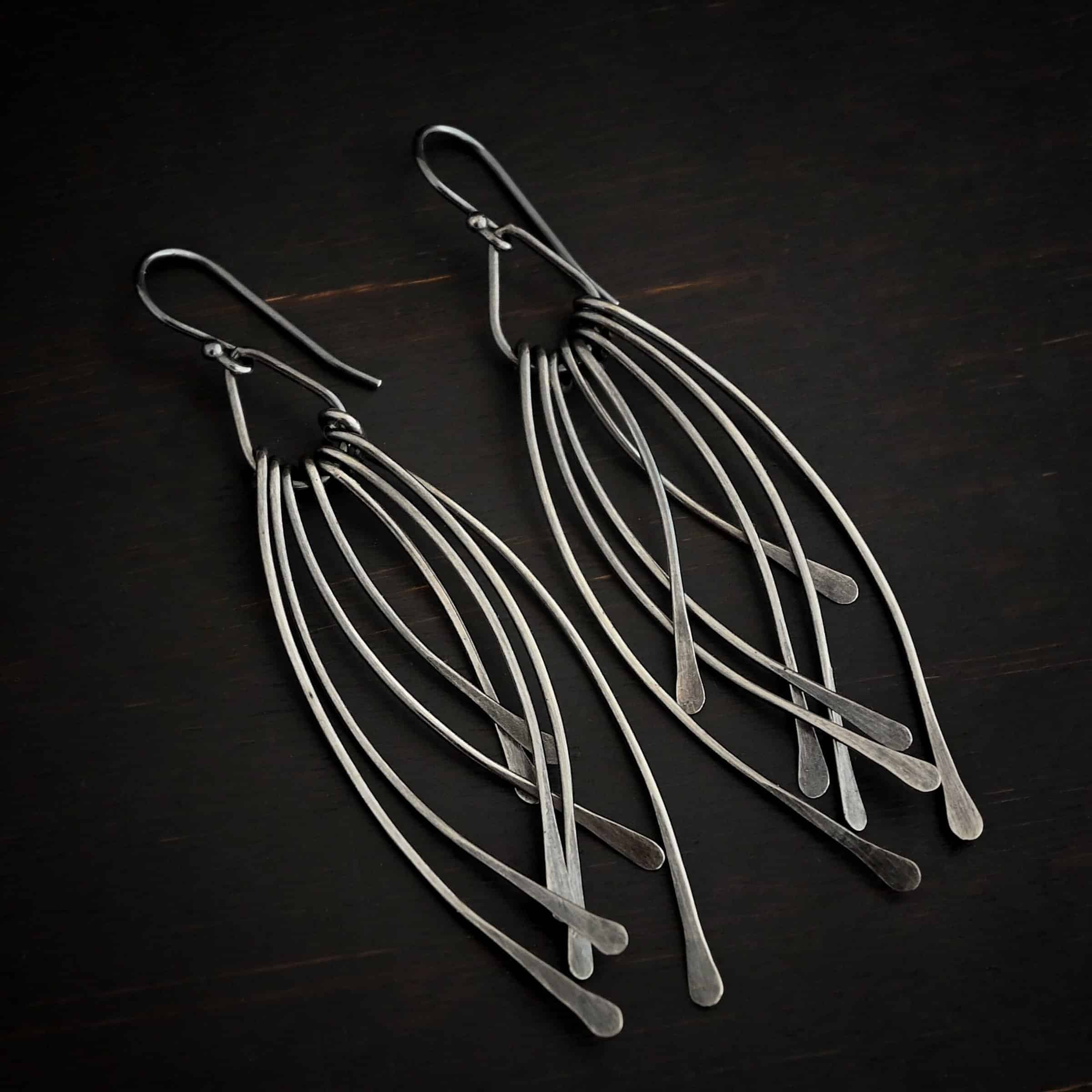 Prairie Wind Earrings Photo of the "Prairie Wind" Earrings. Made out of sterling silver and oxidized. Earrings have french hook ear wires. Attached to the bottom of the ear wires is a teardrop shaped loop of silver. Attached to the bottom of the teardrop loop are eight pieces of silver that all have hammered paddle ends. Four pieces of silver curve towards the right, and the other four pieces curve towards the left, so they overlap each other. Earrings are lying against a dark background.