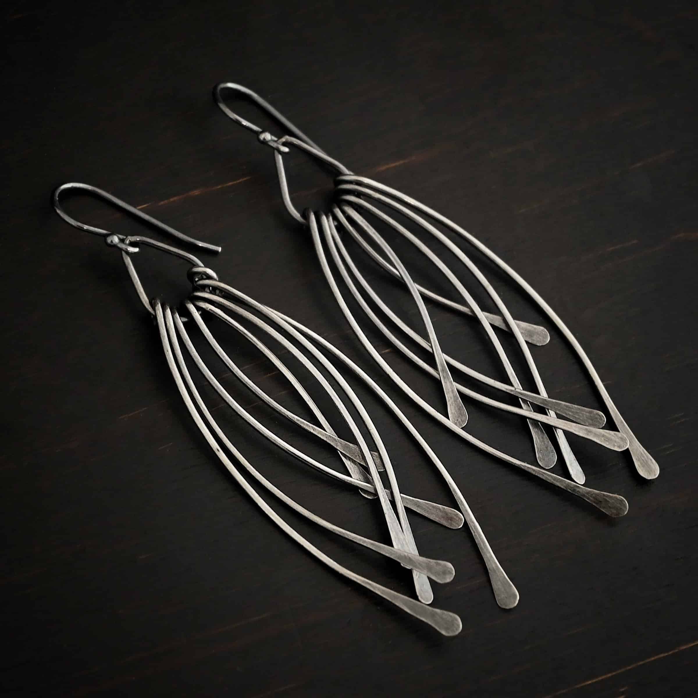 Prairie Wind Earrings Photo of the "Prairie Wind" Earrings. Made out of sterling silver and oxidized. Earrings have french hook ear wires. Attached to the bottom of the ear wires is a teardrop shaped loop of silver. Attached to the bottom of the teardrop loop are eight pieces of silver that all have hammered paddle ends. Four pieces of silver curve towards the right, and the other four pieces curve towards the left, so they overlap each other. Earrings are lying against a dark background.