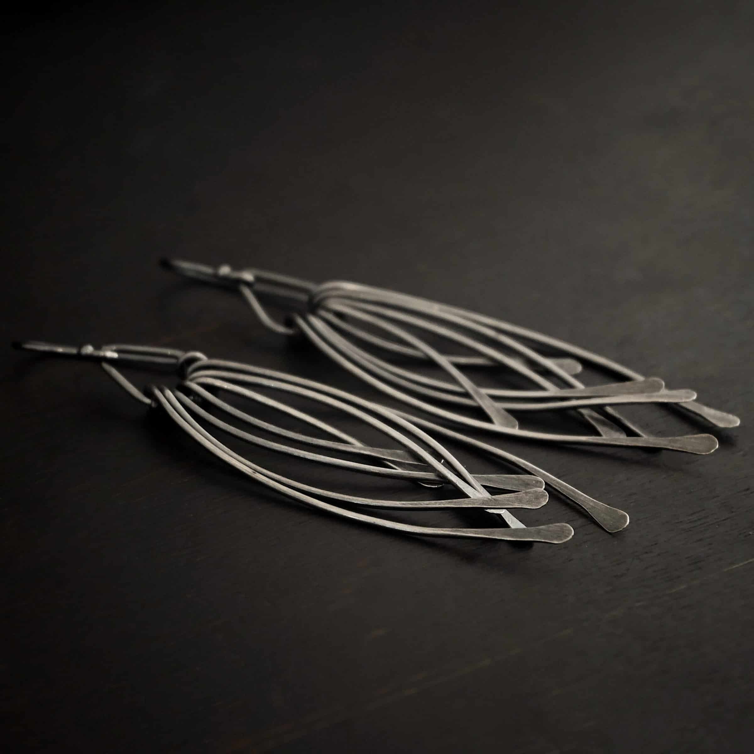 Prairie Wind Earrings Photo of the "Prairie Wind" Earrings. Made out of sterling silver and oxidized. Earrings have french hook ear wires. Attached to the bottom of the ear wires is a teardrop shaped loop of silver. Attached to the bottom of the teardrop loop are eight pieces of silver that all have hammered paddle ends. Four pieces of silver curve towards the right, and the other four pieces curve towards the left, so they overlap each other. Earrings are lying against a dark background.