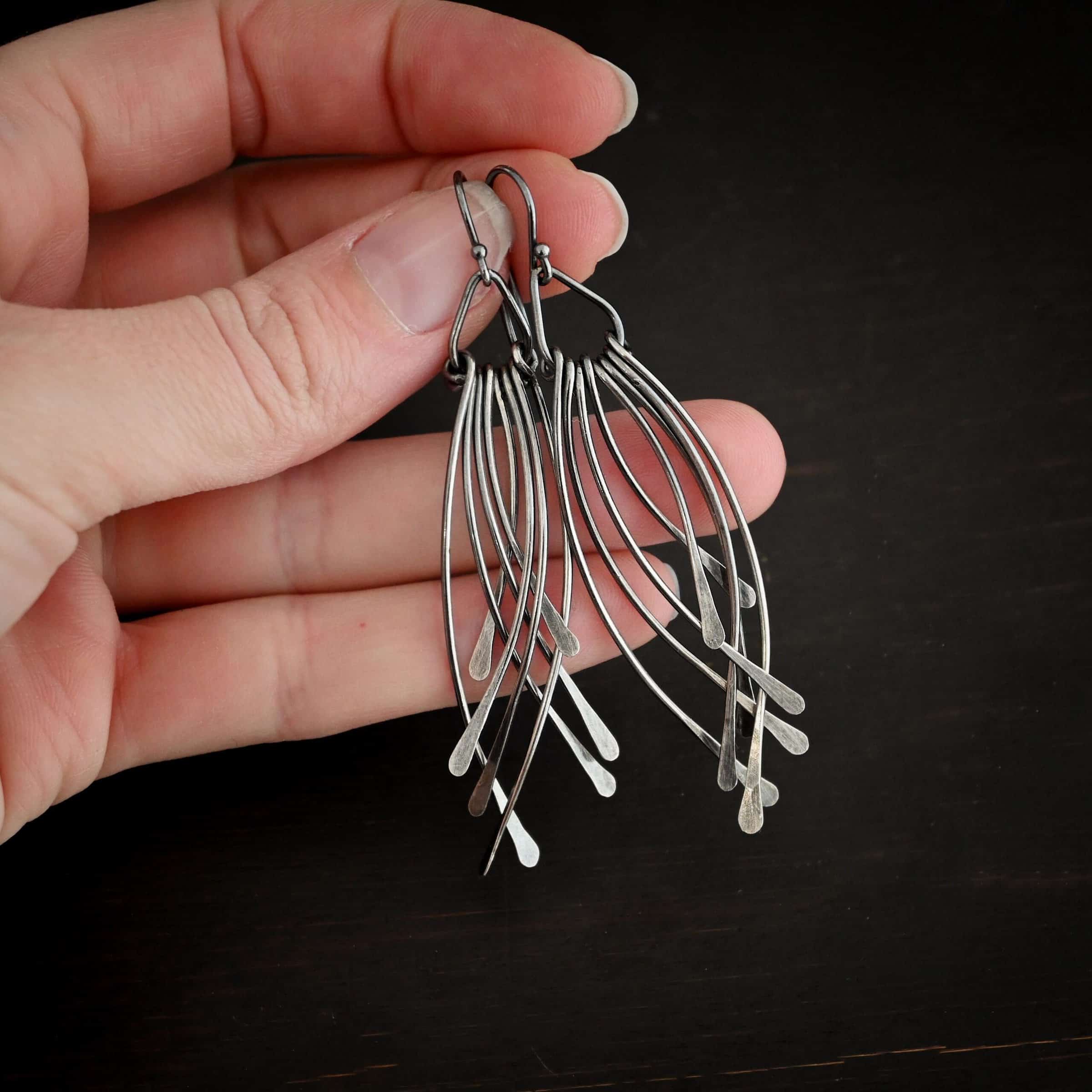 Prairie Wind Earrings Photo of the "Prairie Wind" Earrings. Made out of sterling silver and oxidized. Earrings have french hook ear wires. Attached to the bottom of the ear wires is a teardrop shaped loop of silver. Attached to the bottom of the teardrop loop are eight pieces of silver that all have hammered paddle ends. Four pieces of silver curve towards the right, and the other four pieces curve towards the left, so they overlap each other. Earrings are held in the fingertips of a hand.