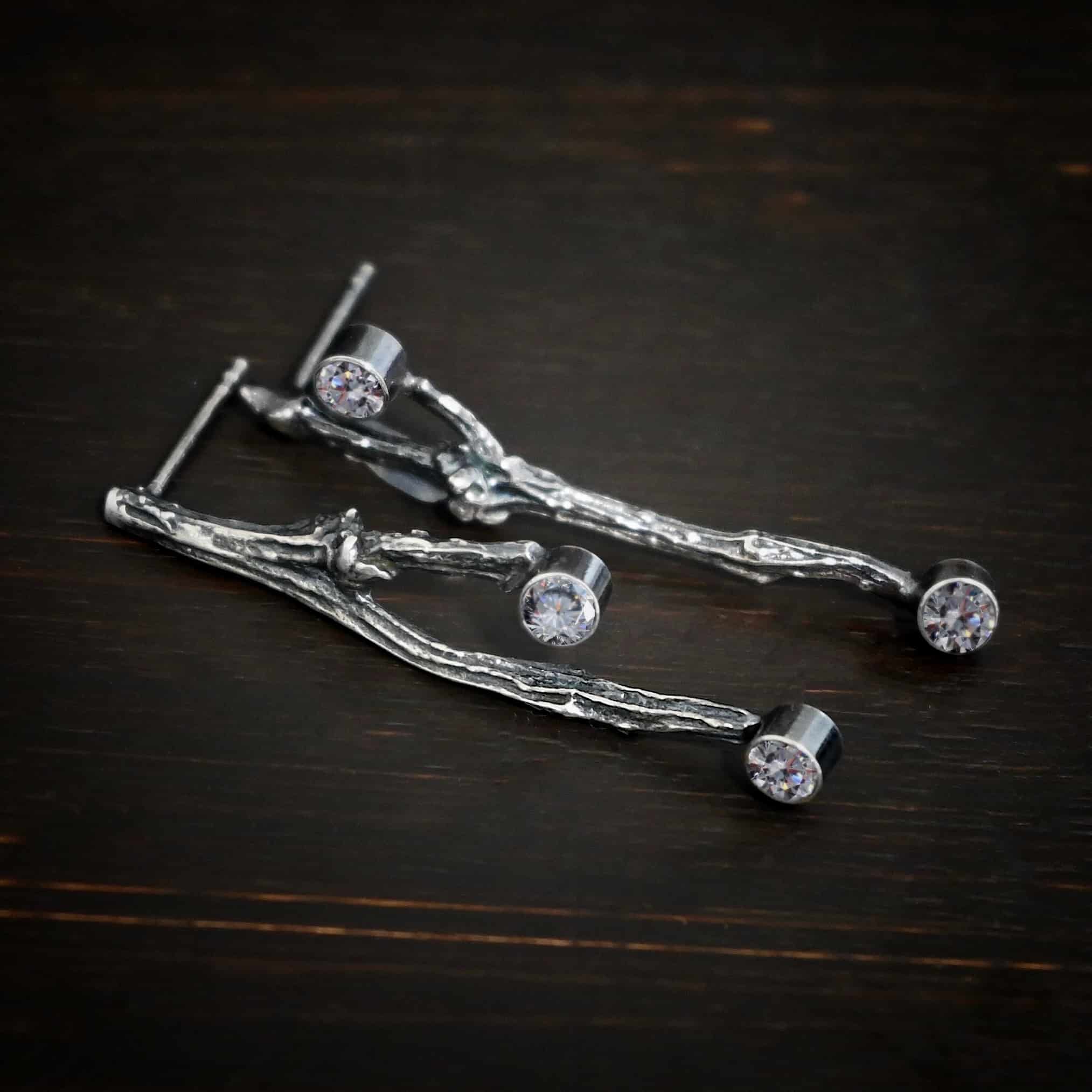 Photo of the "Sticks N Stones" Earrings. Made out of sterling silver and moissanites. The earrings are post earrings and casted sterling silver twigs. The bottom of the twigs each have a small circle moissanite, as well as the top of the right twig and the right side center of the left earring. This means that there are four moissanites total. Earrings are lying against a dark background.