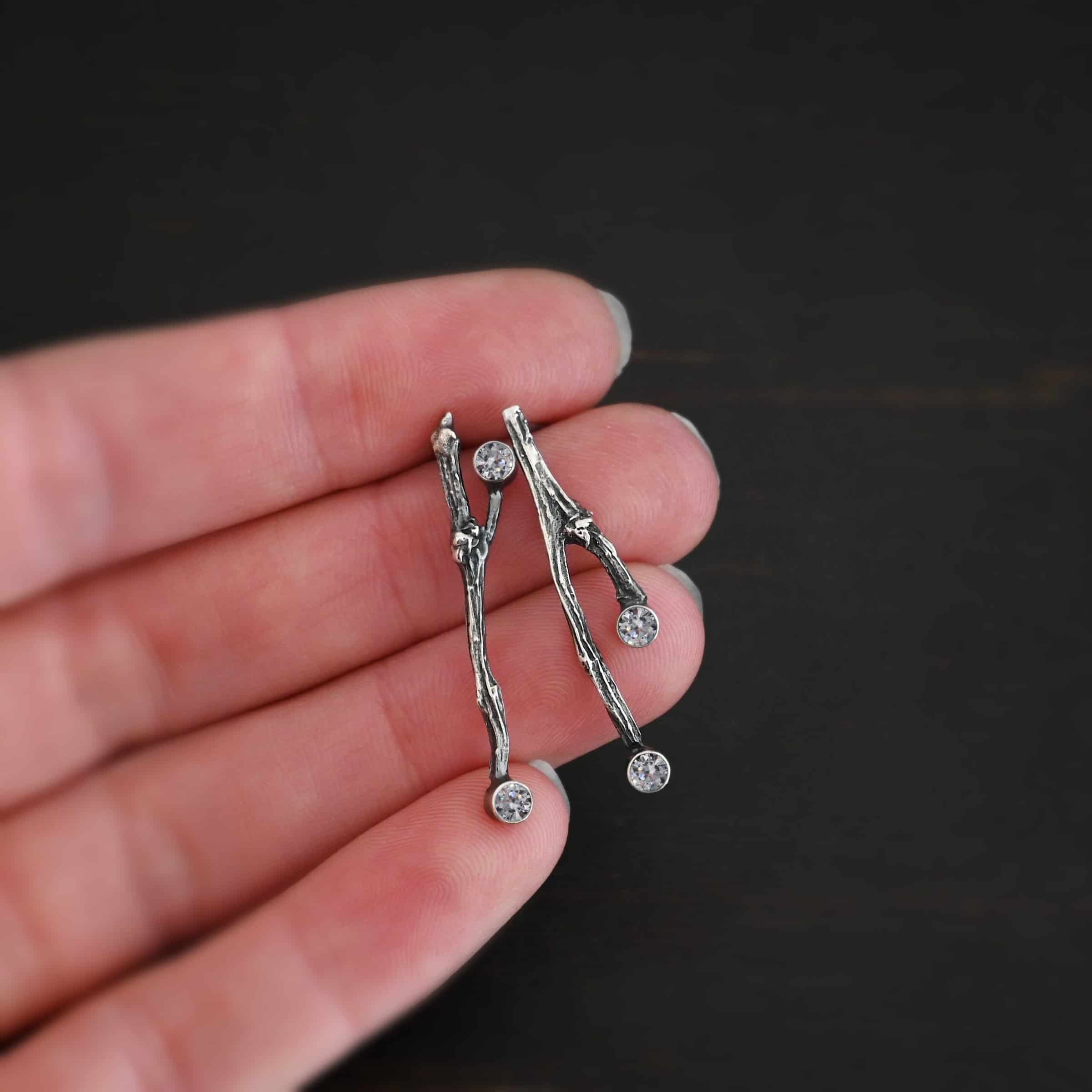 Photo of the "Sticks N Stones" Earrings. Made out of sterling silver and moissanites. The earrings are post earrings and casted sterling silver twigs. The bottom of the twigs each have a small circle moissanite, as well as the top of the right twig and the right side center of the left earring. This means that there are four moissanites total. Earrings are held in the fingertips of a hand.