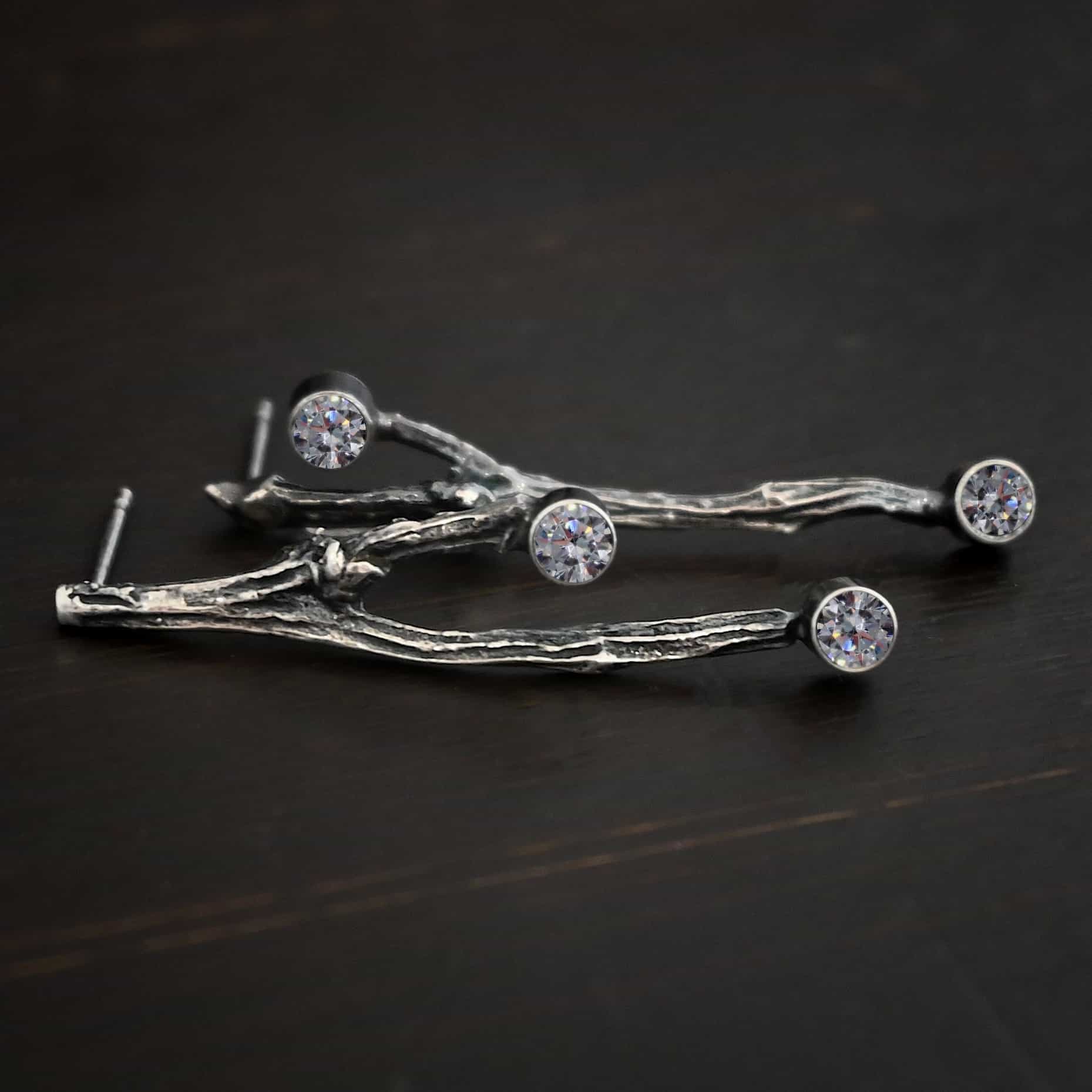 Photo of the "Sticks N Stones" Earrings. Made out of sterling silver and moissanites. The earrings are post earrings and casted sterling silver twigs. The bottom of the twigs each have a small circle moissanite, as well as the top of the right twig and the right side center of the left earring. This means that there are four moissanites total. Earrings are lying against a dark background.